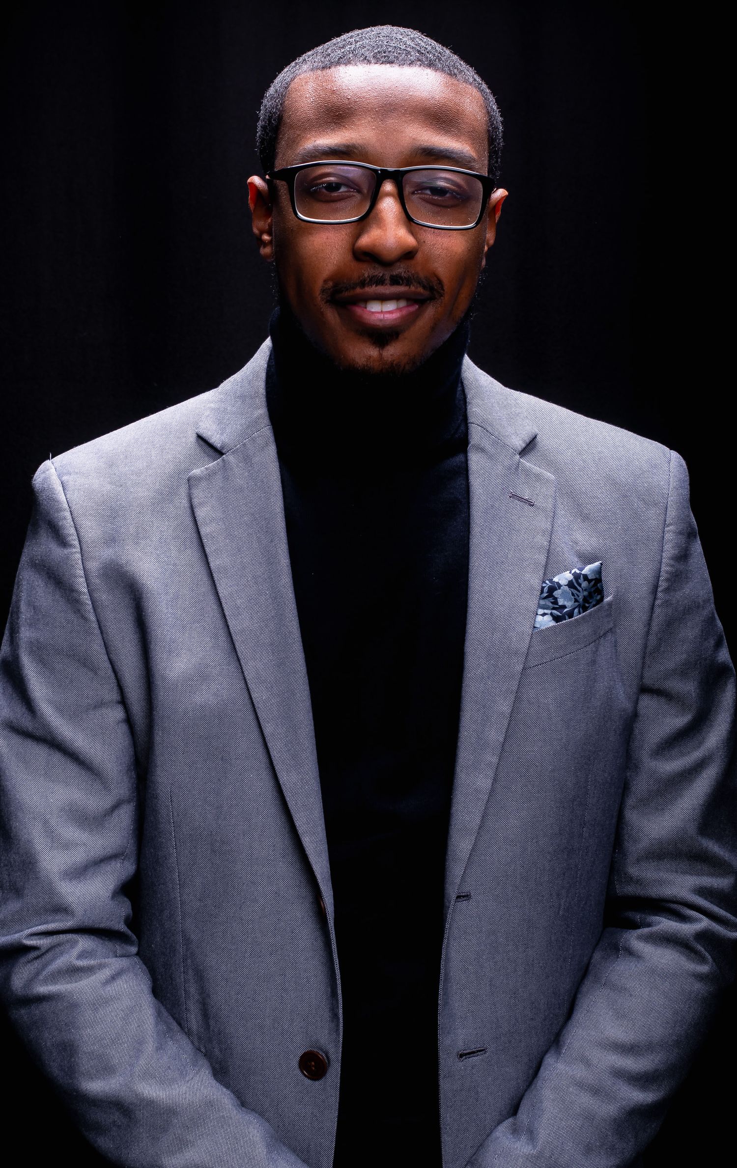 Confident Black man in gray blazer and black turtleneck with glasses on dark background.
