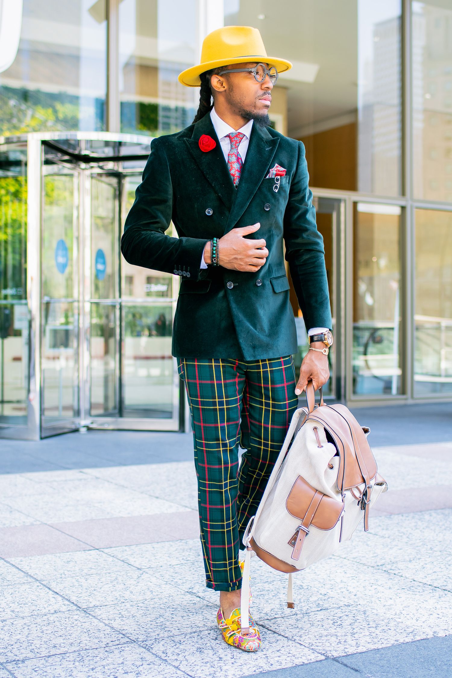Fashionable man in yellow fedora, dark green blazer, plaid trousers, carrying pink backpack.