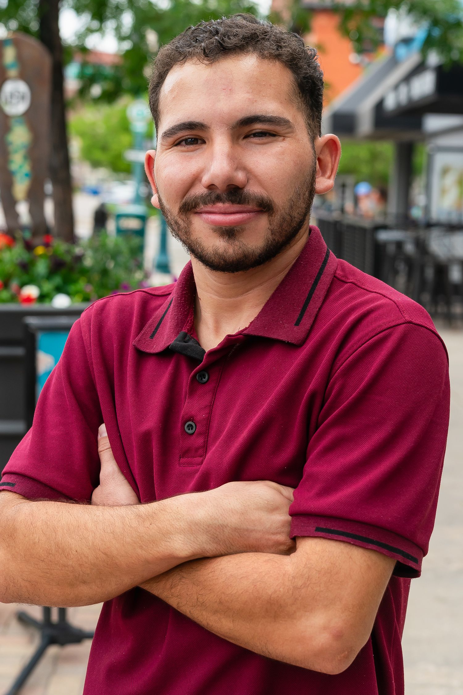 Young Hispanic man with beard in maroon polo shirt smiling with arms crossed on urban street.