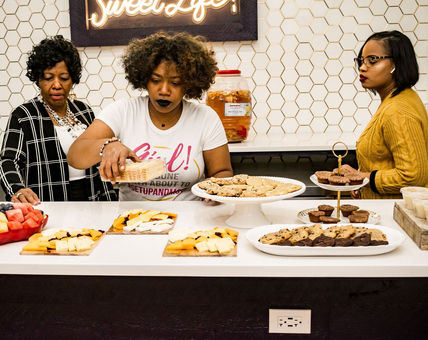 Three women arranging assorted pastries and desserts on a white counter inside a bakery.