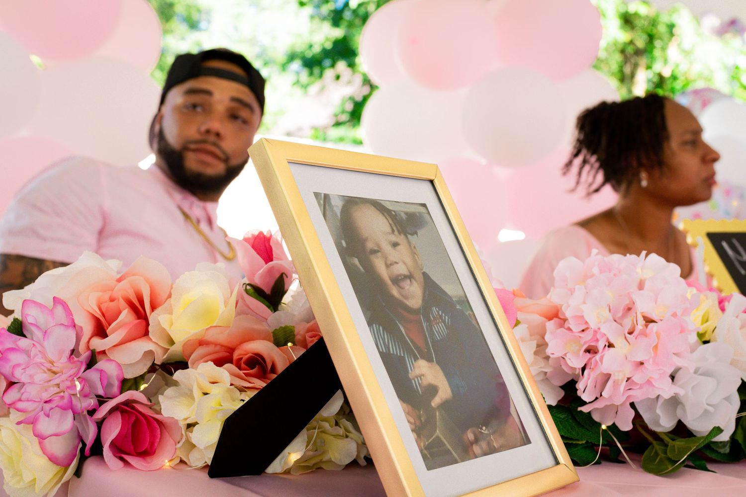 A framed photo displayed among pink flowers at a baby shower celebration with guests in background.