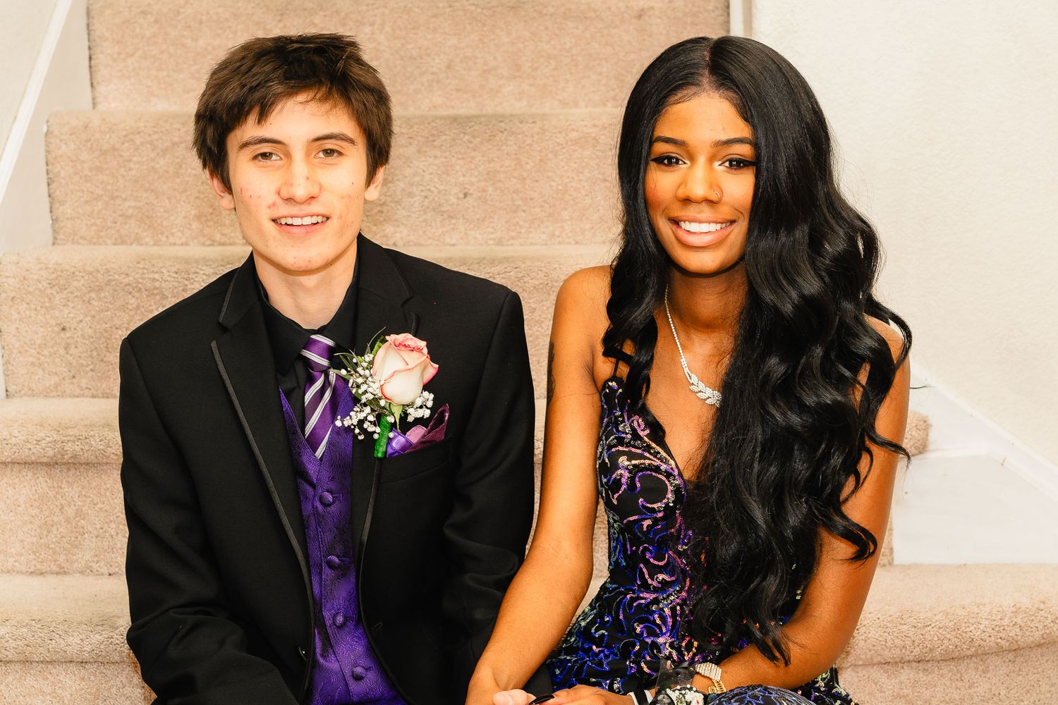Teen couple dressed formally in prom attire sitting on stairs, boy in black tuxedo and girl in blue sequin dress.