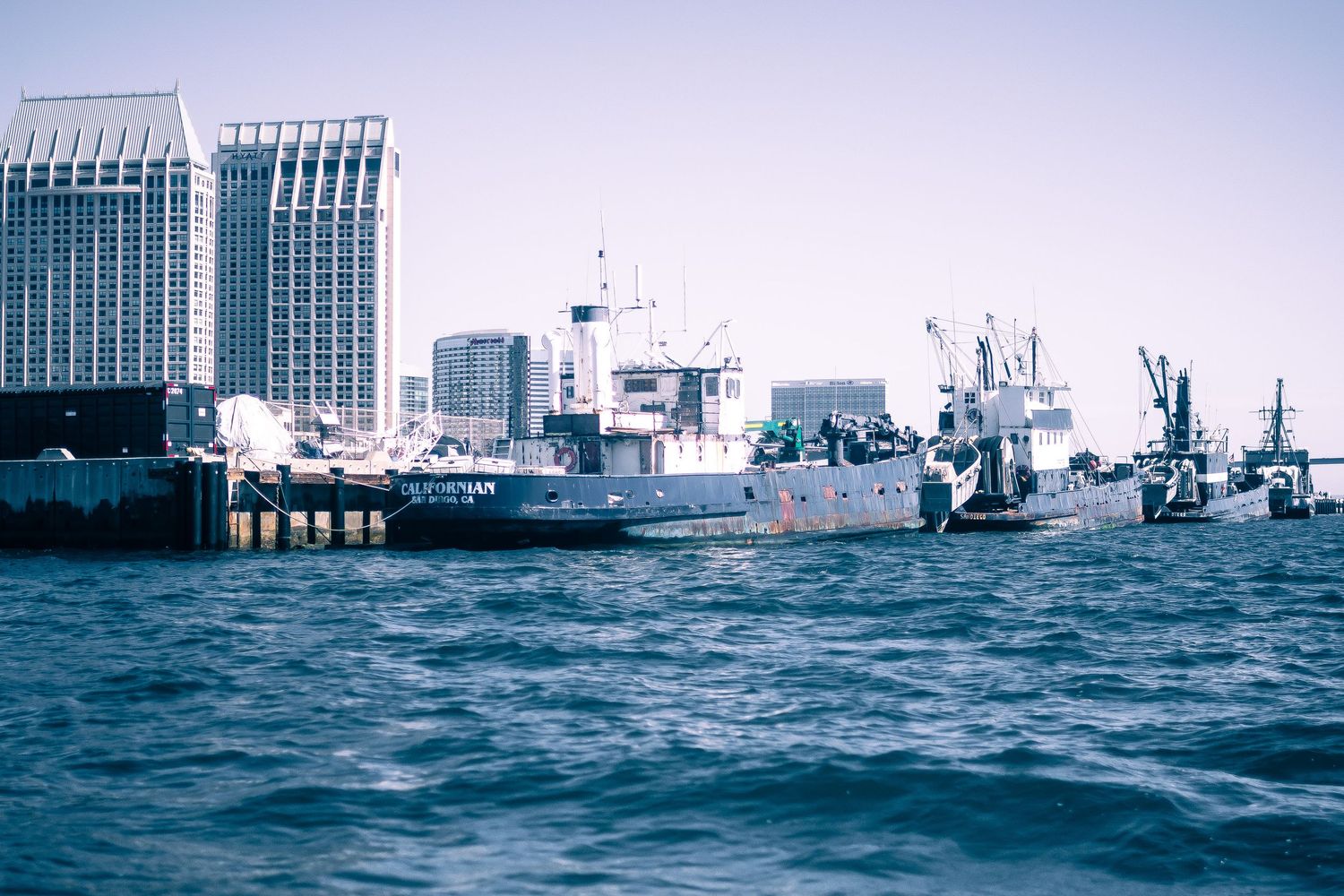 Military vessels and ships docked at a harbor with city skyline in the background.