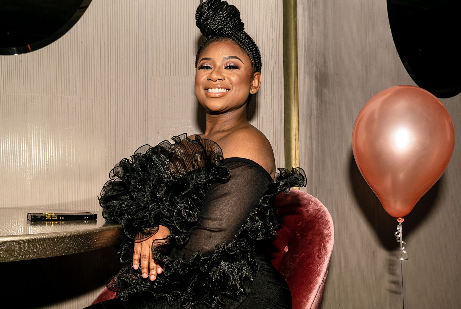 Smiling woman in a black ruffled off-shoulder dress seated near a pink balloon at a party.