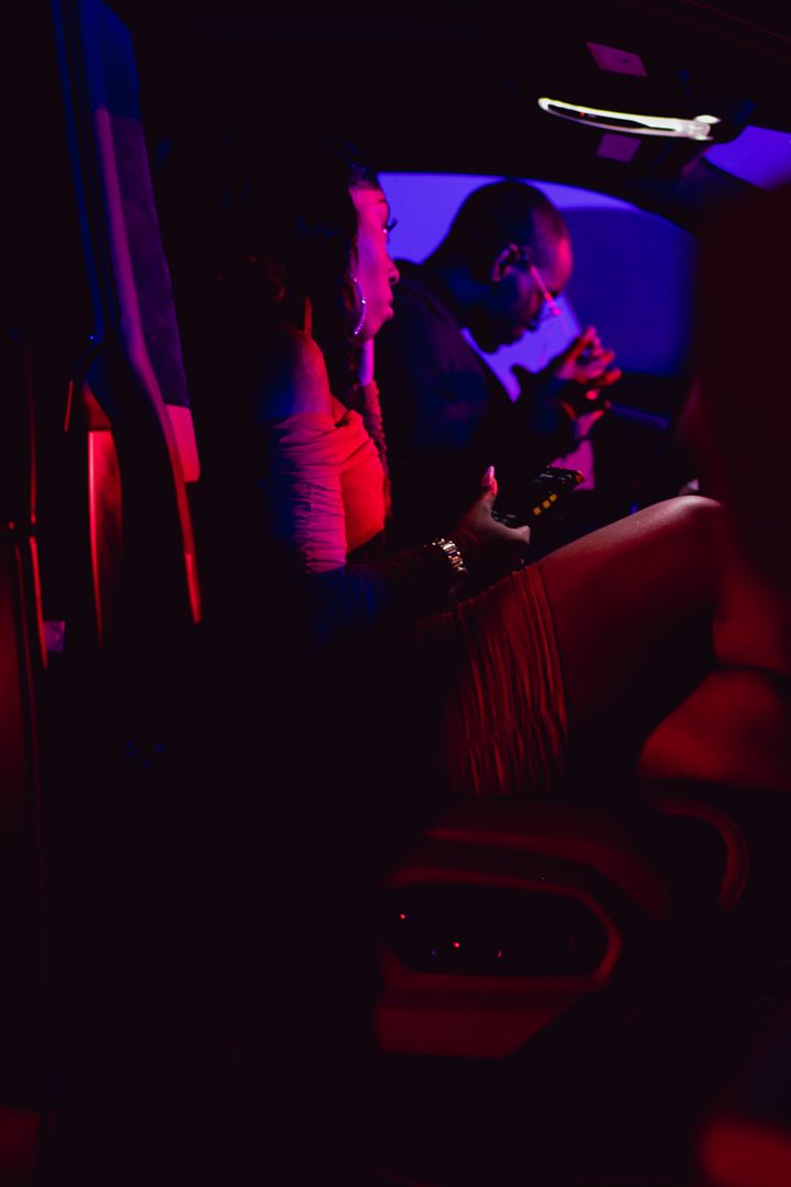 Two people sit in a luxury car with red leather interior under dim purple lighting.