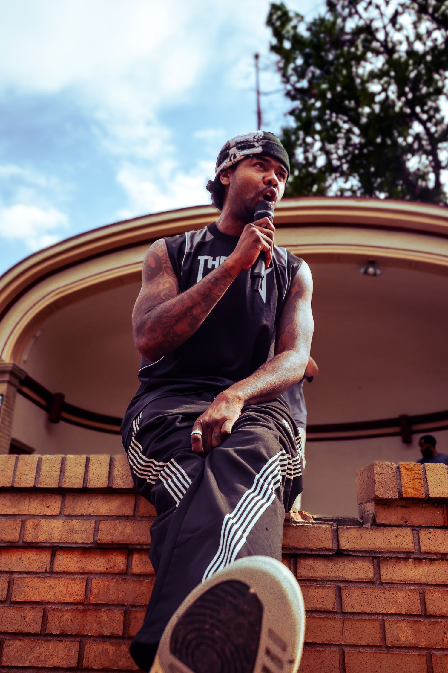 Athletic Black man in sleeveless shirt and bandana sitting on brick wall outdoors under blue sky.