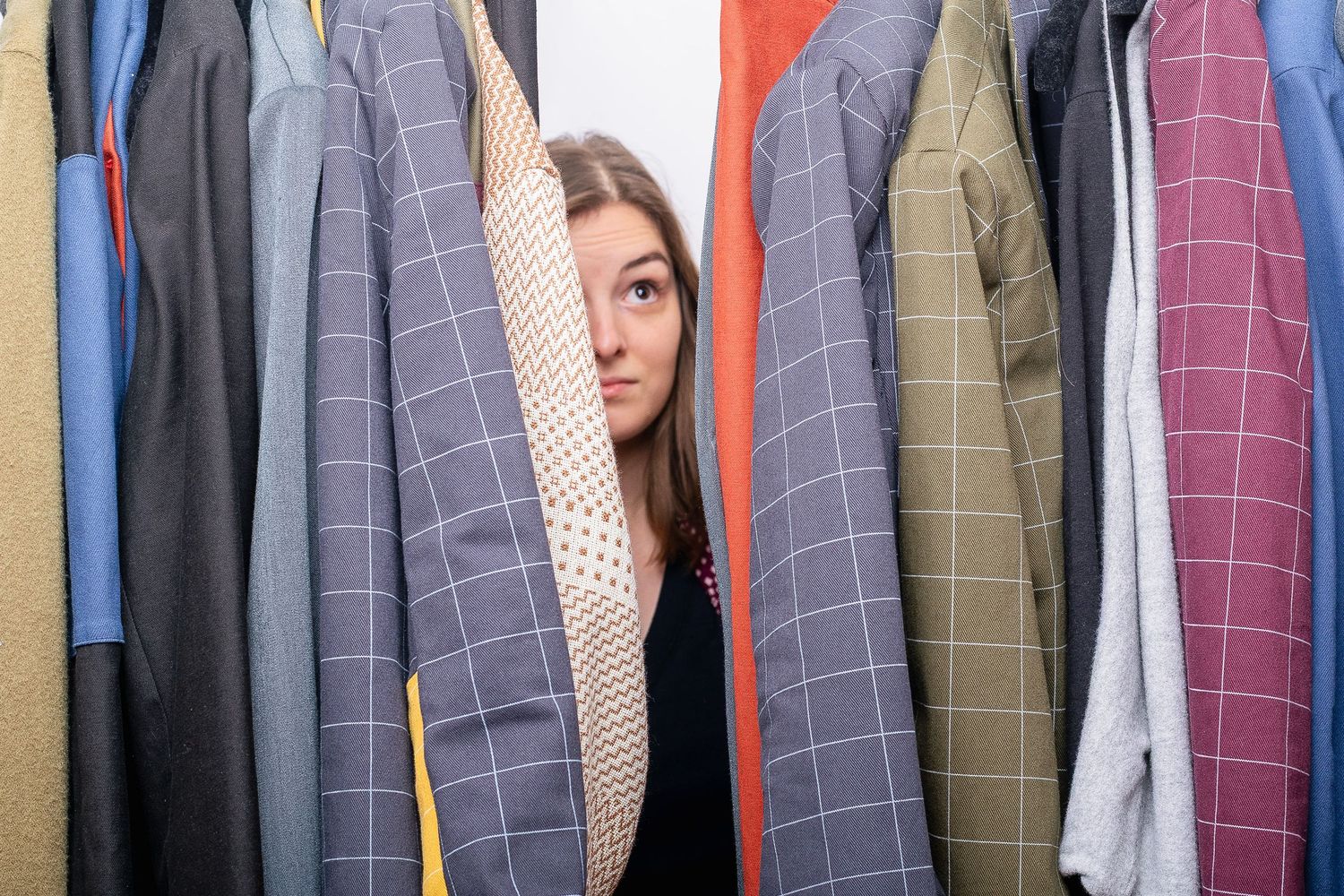 Woman peeking through colorful hanging jackets in a closet full of patterned blazers.