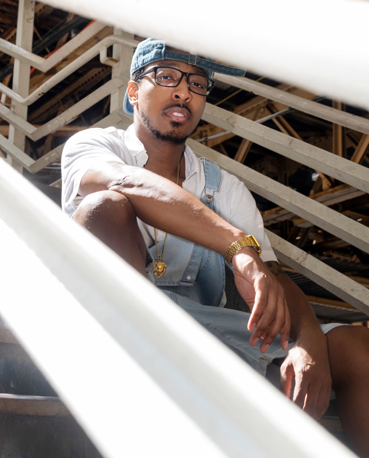 Young man in denim overalls and gold watch sits on metal stairs outdoors.