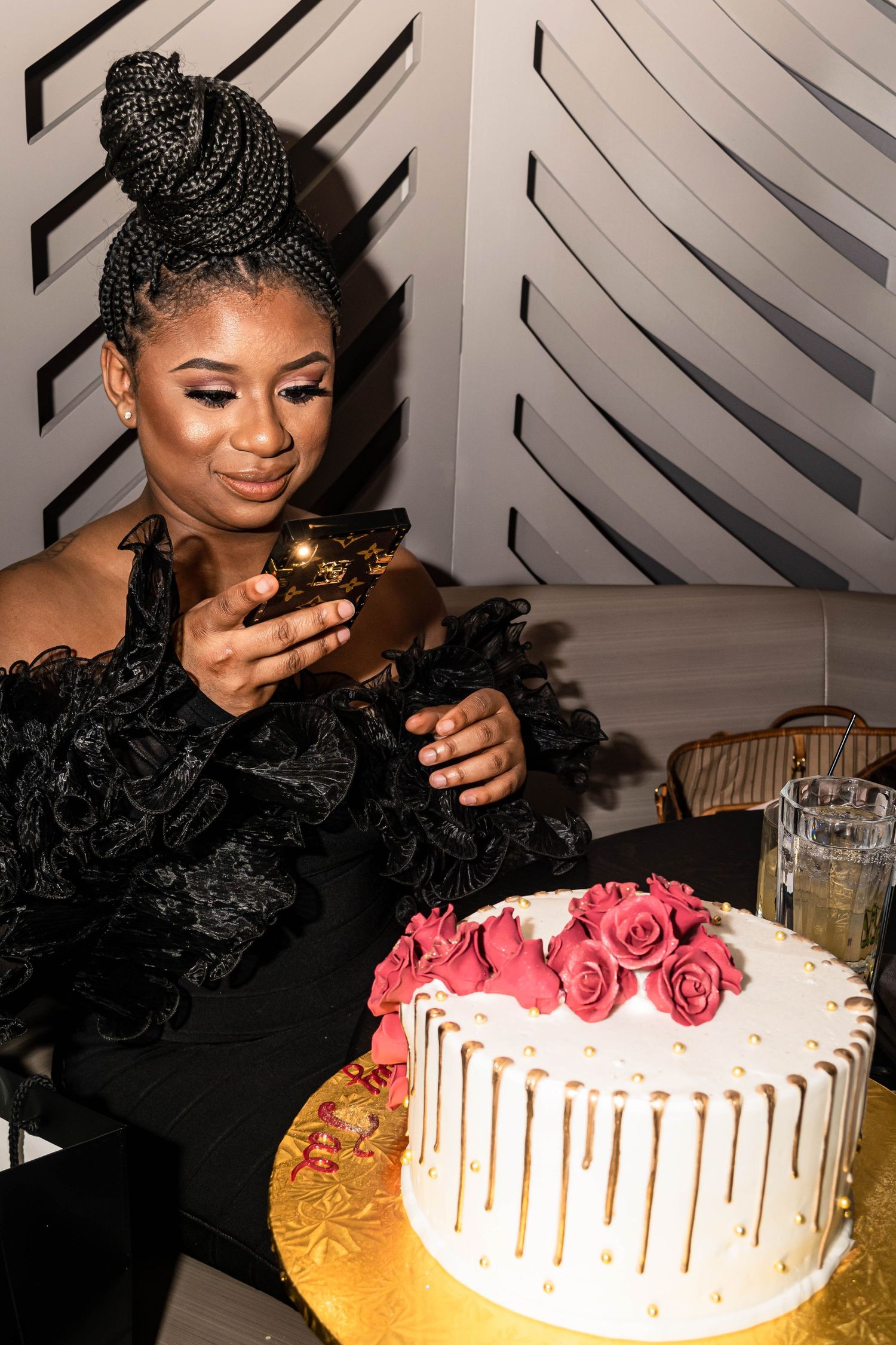 Elegant woman in black ruffled dress admiring a white cake decorated with pink roses and gold drips.