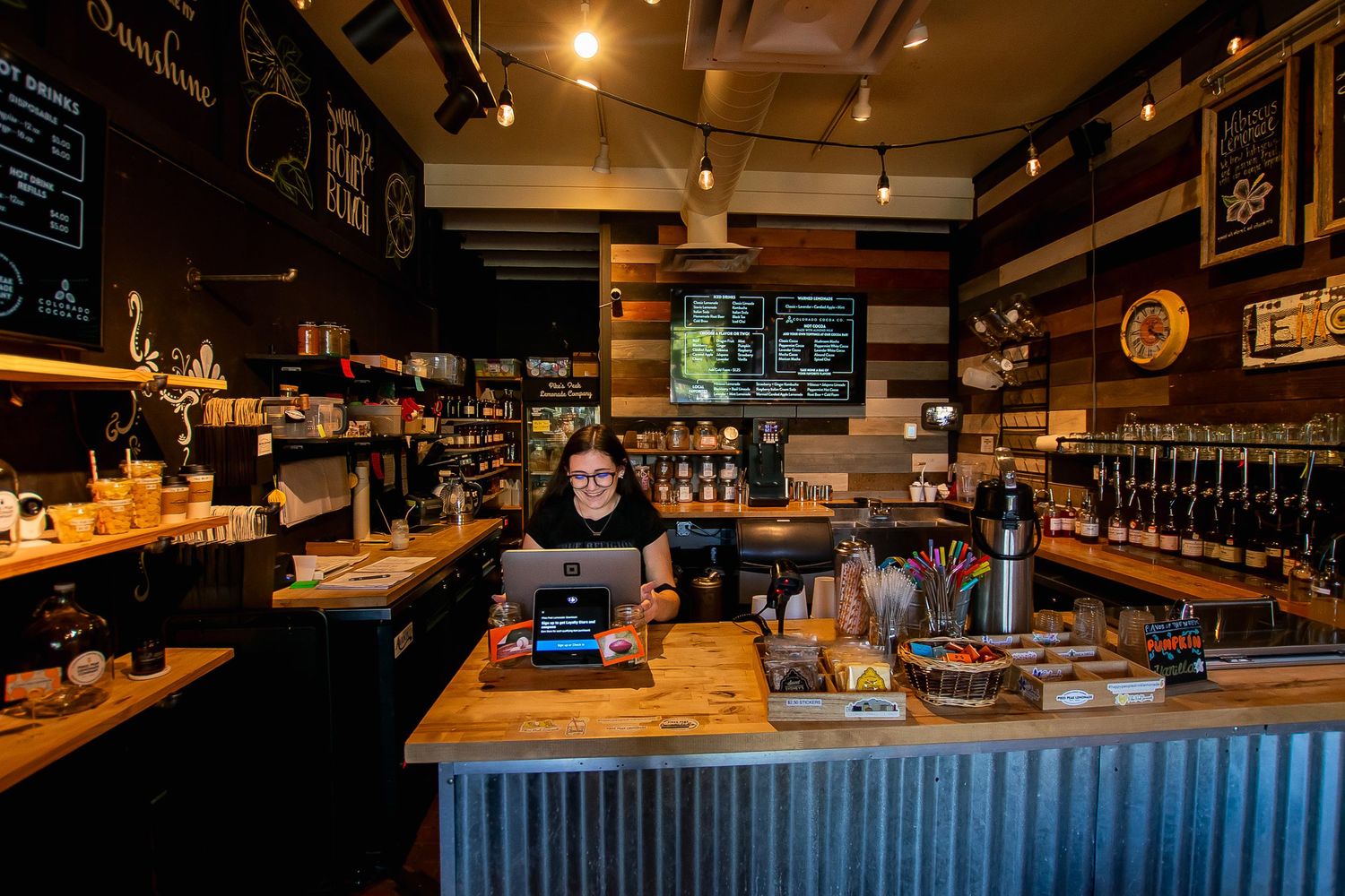 A female barista works at a laptop behind a rustic wooden bar in a cozy cafe.