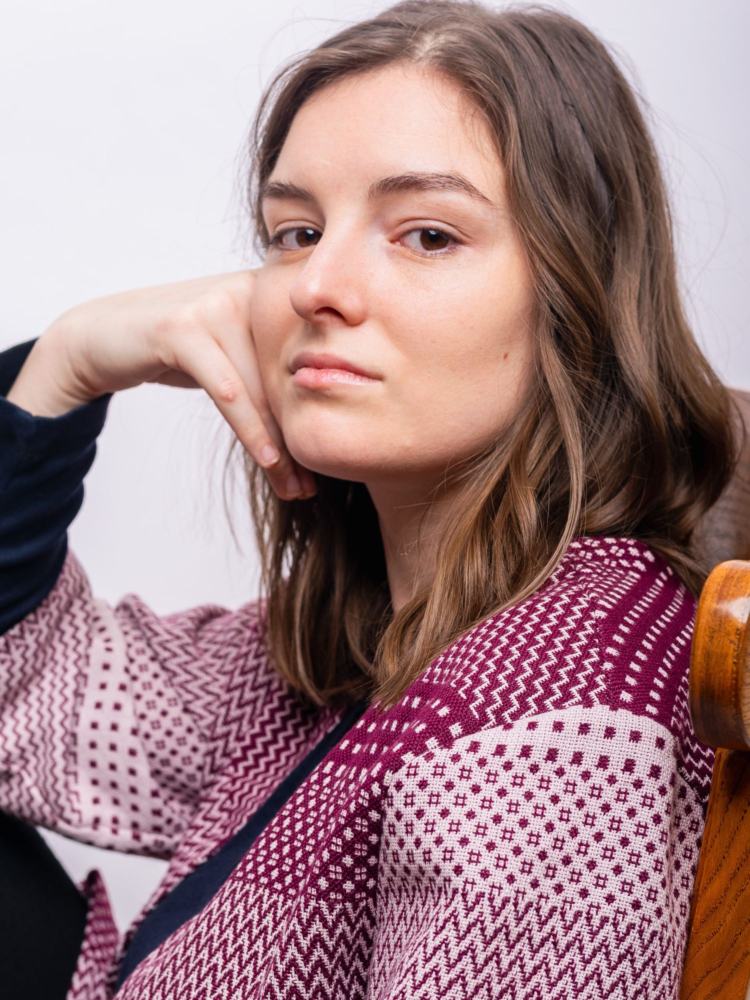 Young woman in a burgundy patterned cardigan resting her chin on her hand while seated in a wooden chair.