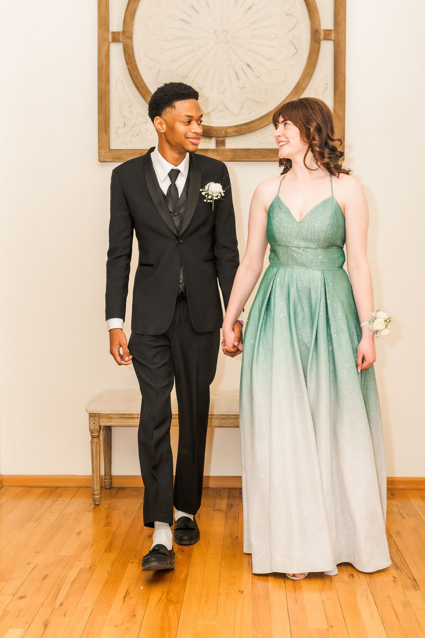 Young interracial couple in formal prom attire holding hands, boy in black tuxedo, girl in green ombre gown.