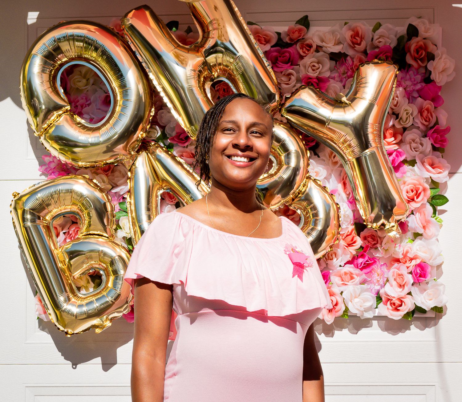 Smiling pregnant Black woman in pink dress standing before OH BABY gold balloon letters and floral wall.