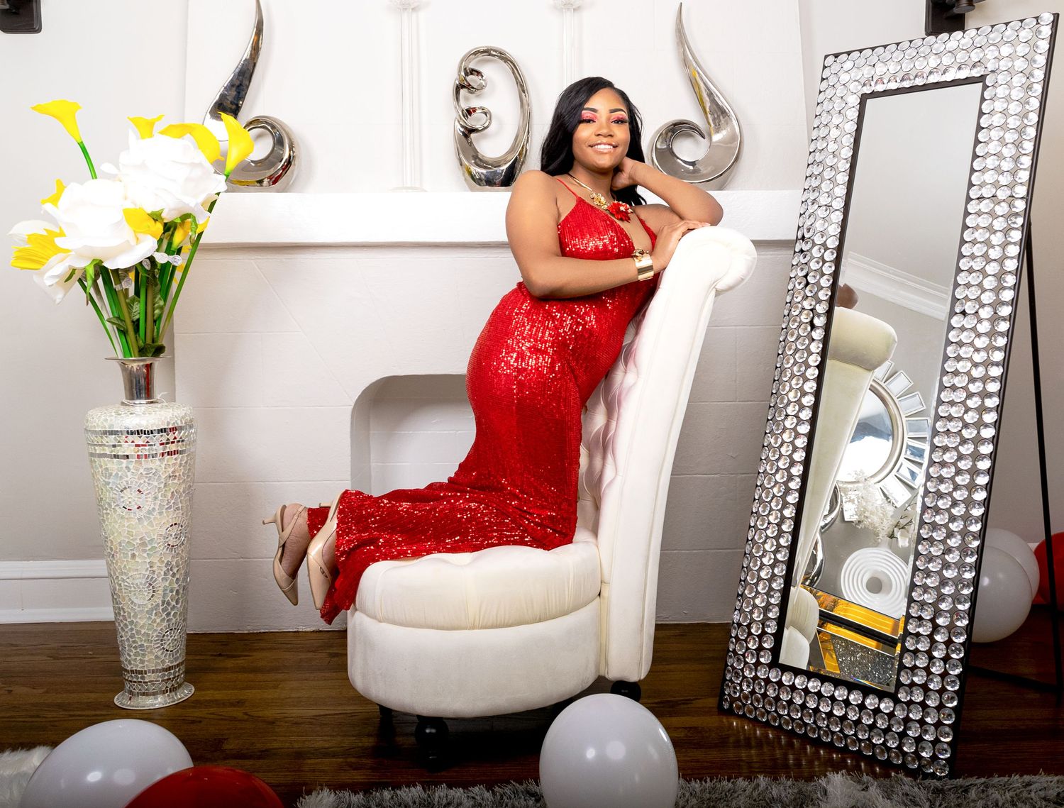 Woman in a red sequin gown sitting gracefully in a white chair beside a decorative mirror.