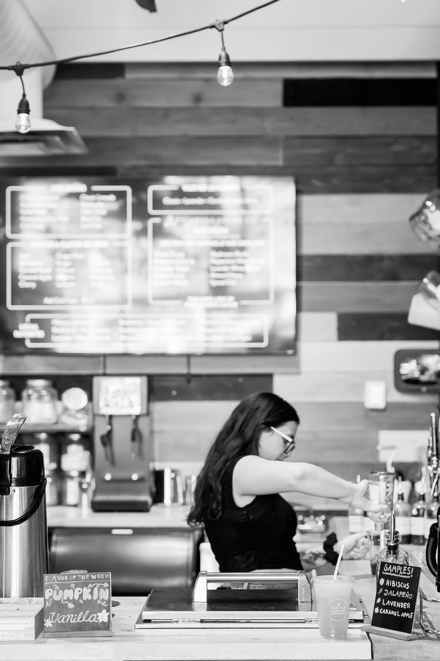 Black and white photo of female barista working behind coffee shop counter with menu board behind her.