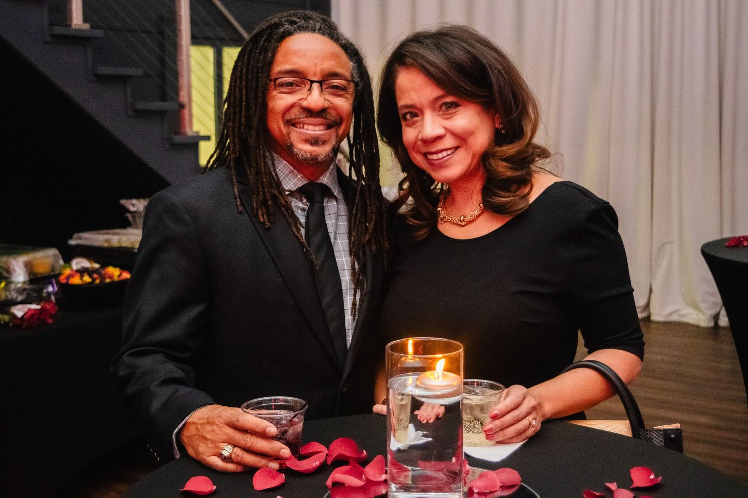 Smiling couple holds drinks at an elegant candlelit event with rose petals on table.