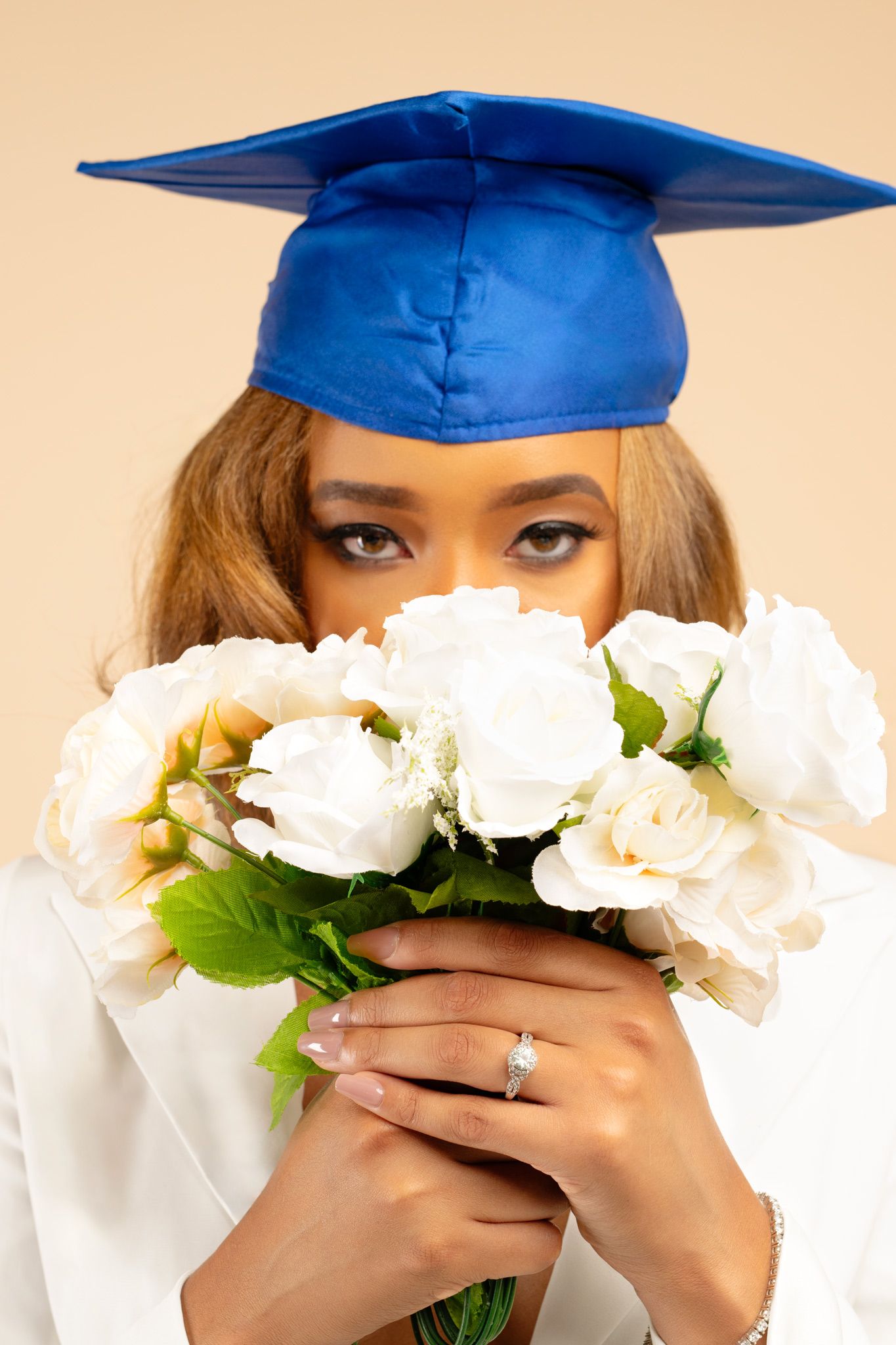 Graduate in blue cap holds white rose bouquet covering face, wearing diamond ring on beige background.