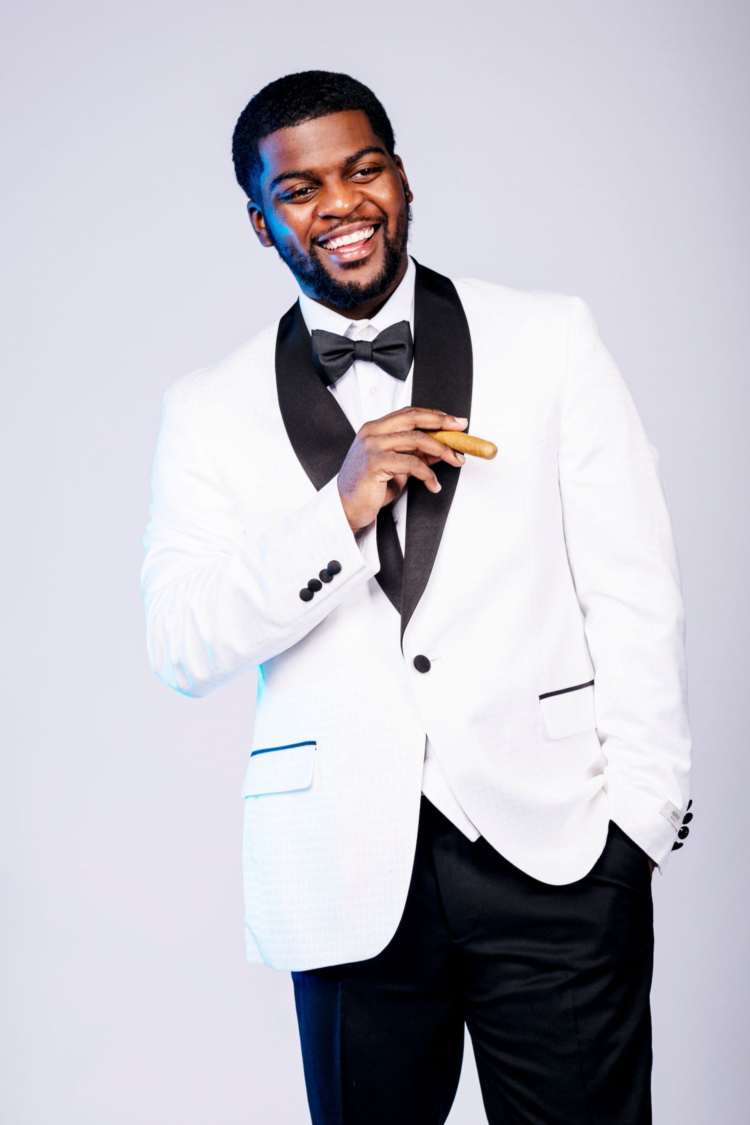 Smiling man in a white tuxedo jacket with black bow tie holding a cigar confidently.