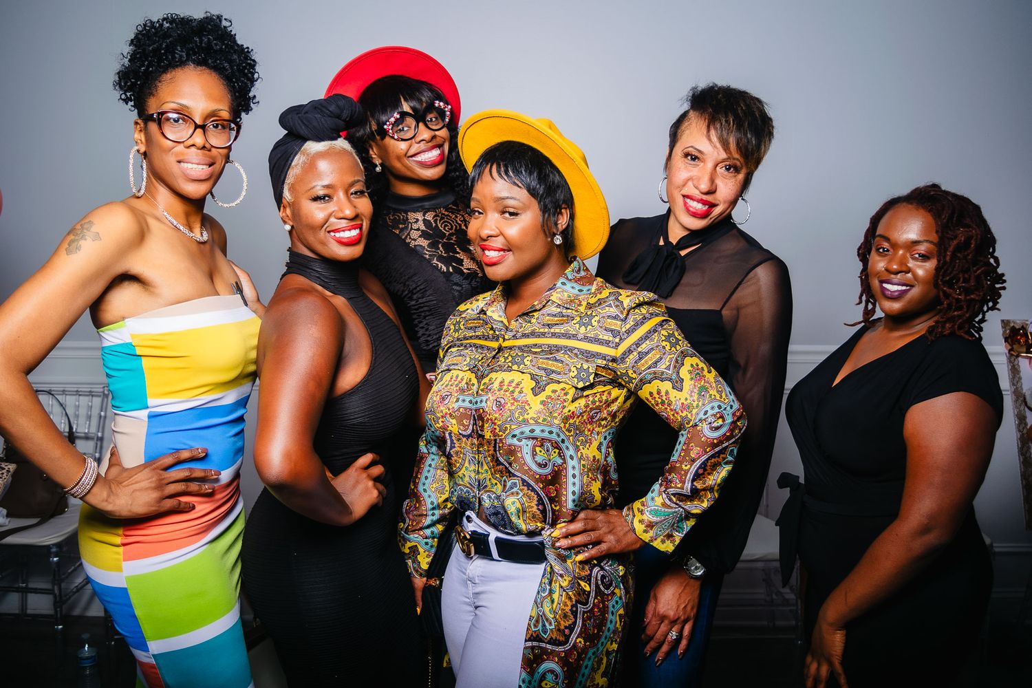 Group of six women in colorful outfits smile together at a lively social gathering.
