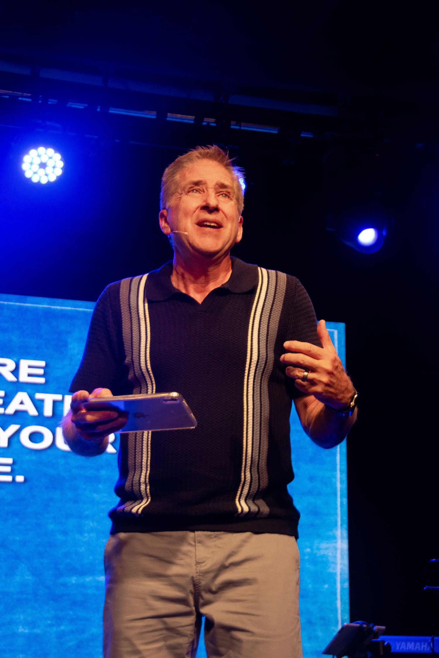 Senior man in striped polo shirt speaking on stage holding tablet under blue stage lighting.