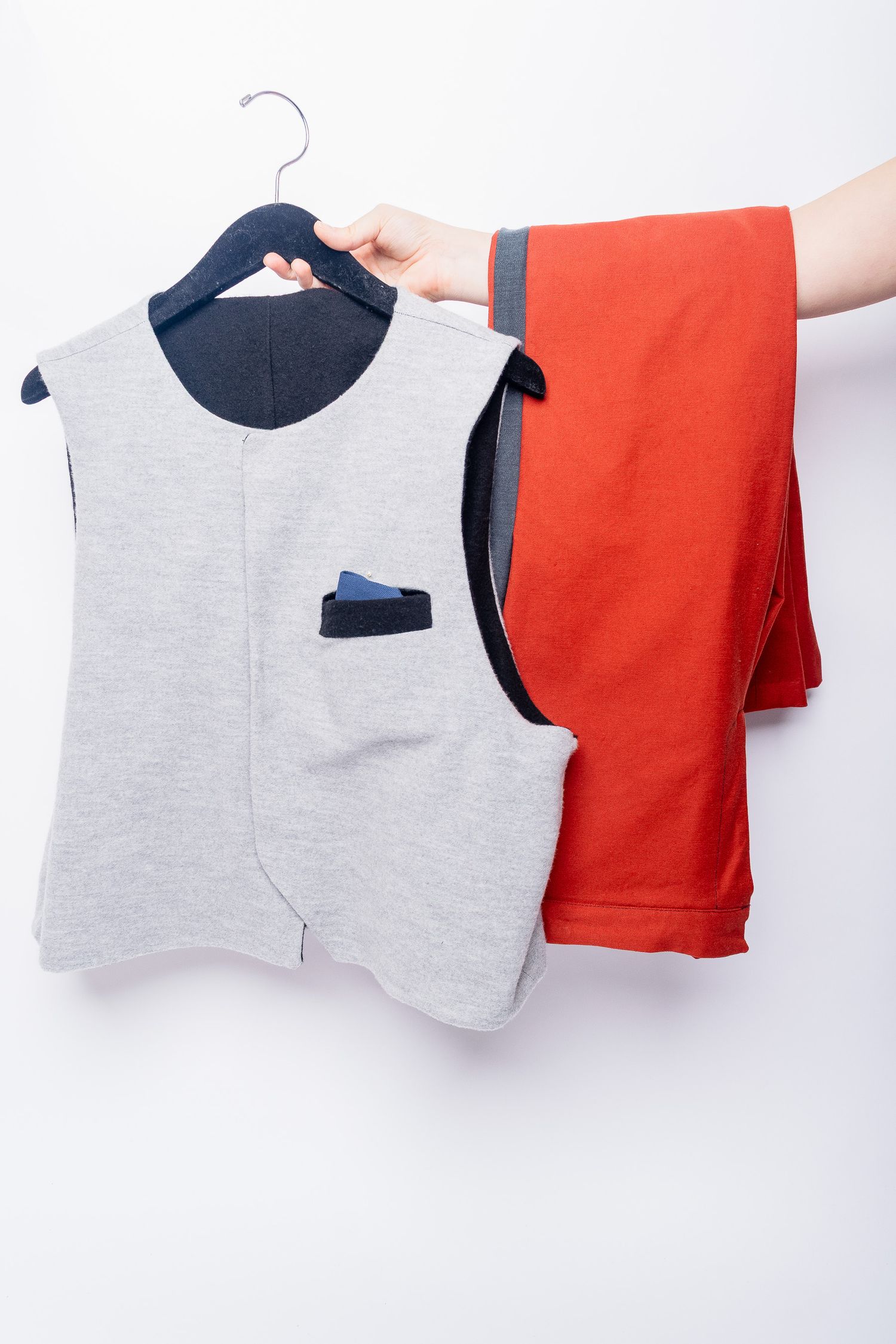 Hand holding a gray sleeveless vest on hanger next to folded red pants on white background.