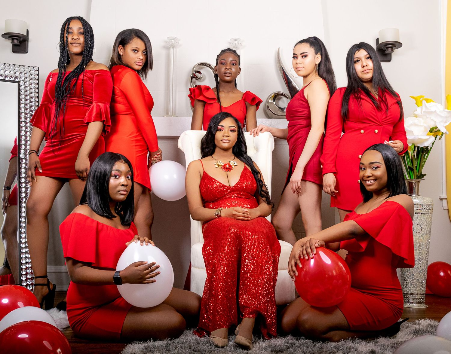 Group of eight women dressed in red outfits posing together with balloons in a decorated room.