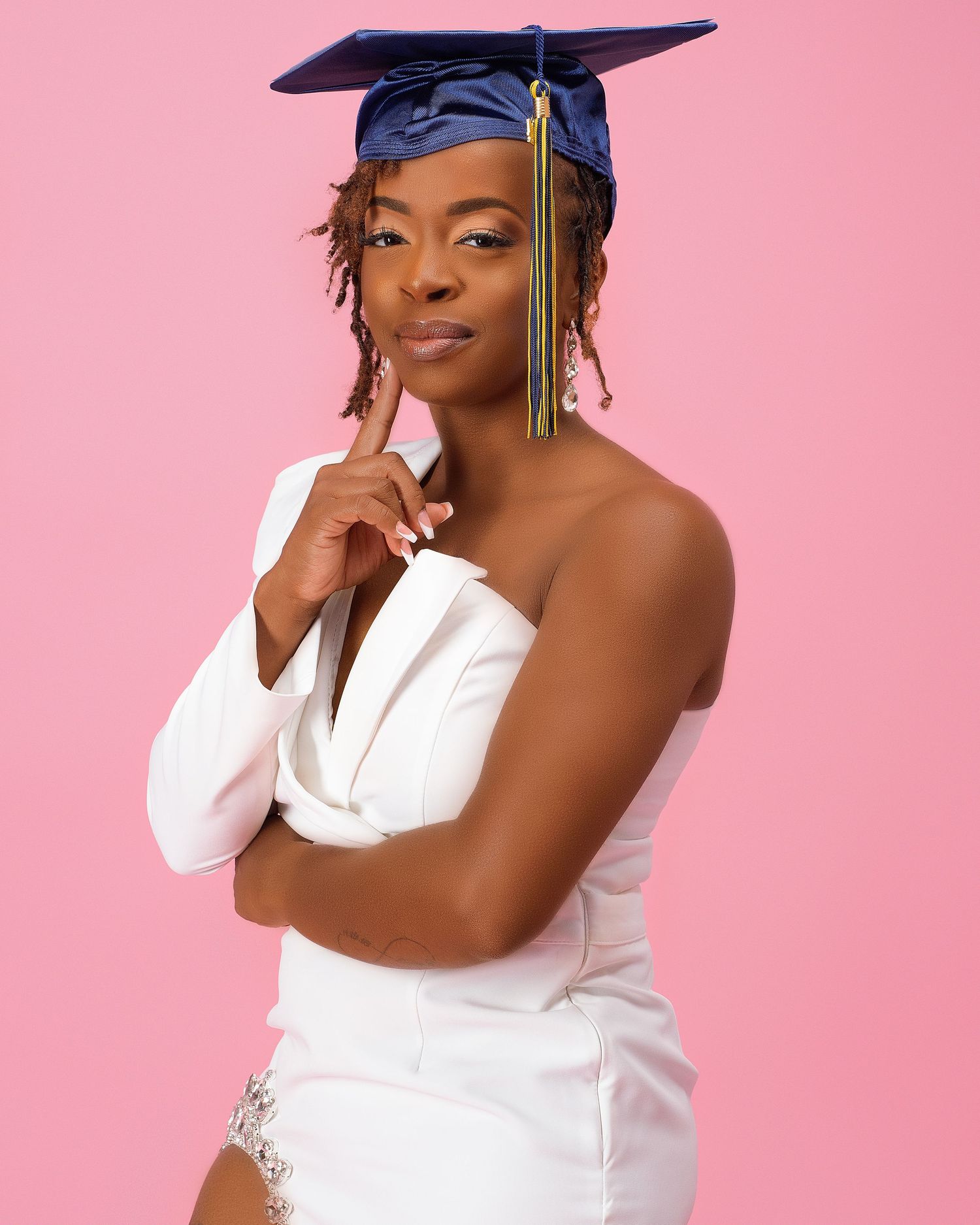 Young Black woman in white strapless outfit and blue graduation cap poses thoughtfully against pink background.