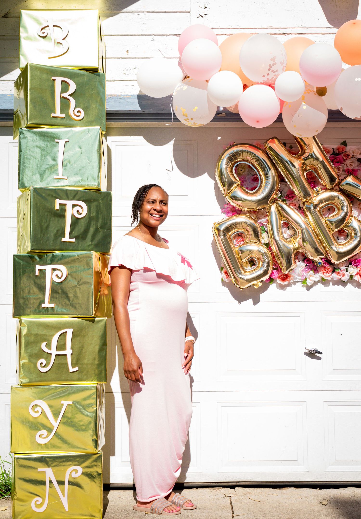 Pregnant woman in pink dress standing beside letter blocks spelling BRITAYN at an outdoor baby shower.