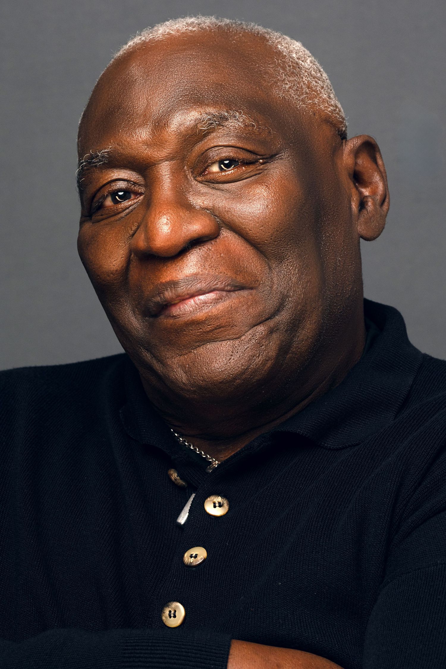 Smiling elderly Black man with gray hair wearing a black polo shirt with arms crossed.