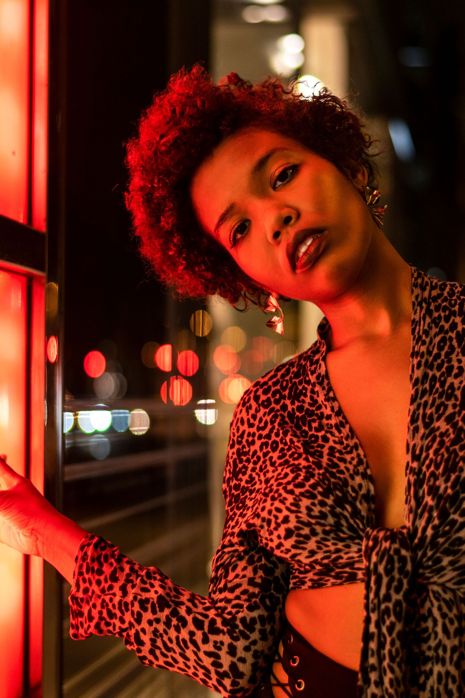 Nighttime portrait of a woman with red curly hair in a leopard print top near neon lights.