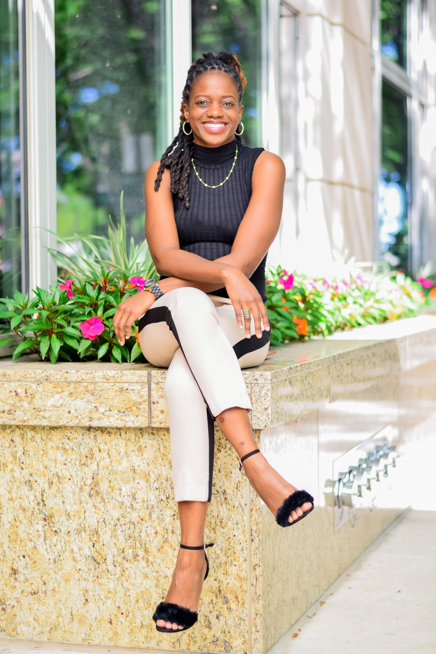 Smiling woman in black sleeveless top and beige pants sitting on stone ledge with flowers outdoors.