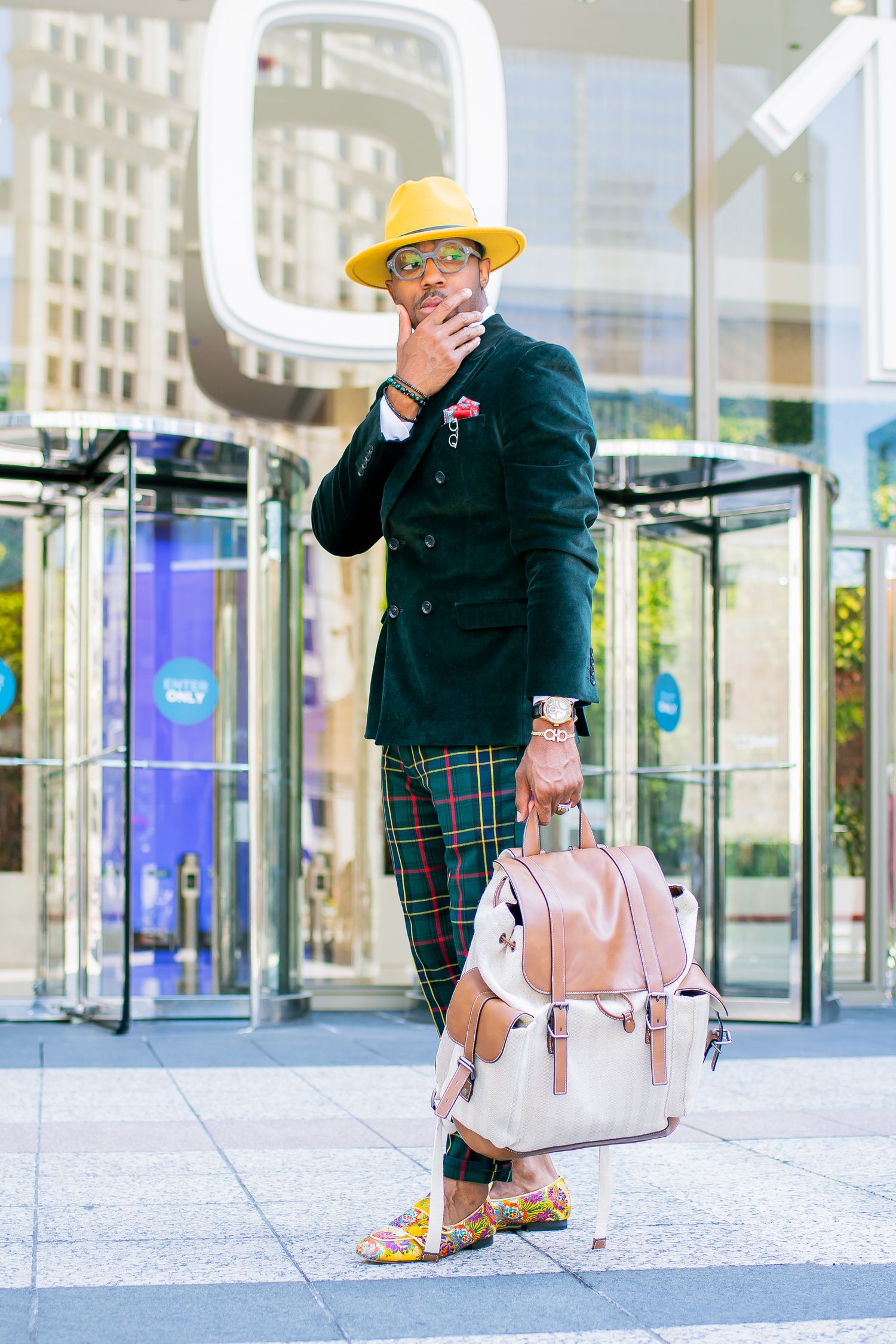 Stylish man in yellow hat, green blazer, plaid pants, holding pink backpack outdoors.