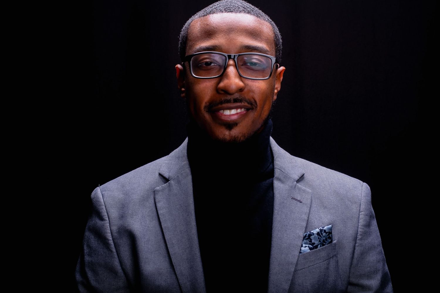 Smiling Black man in gray blazer and black turtleneck with glasses against dark background.