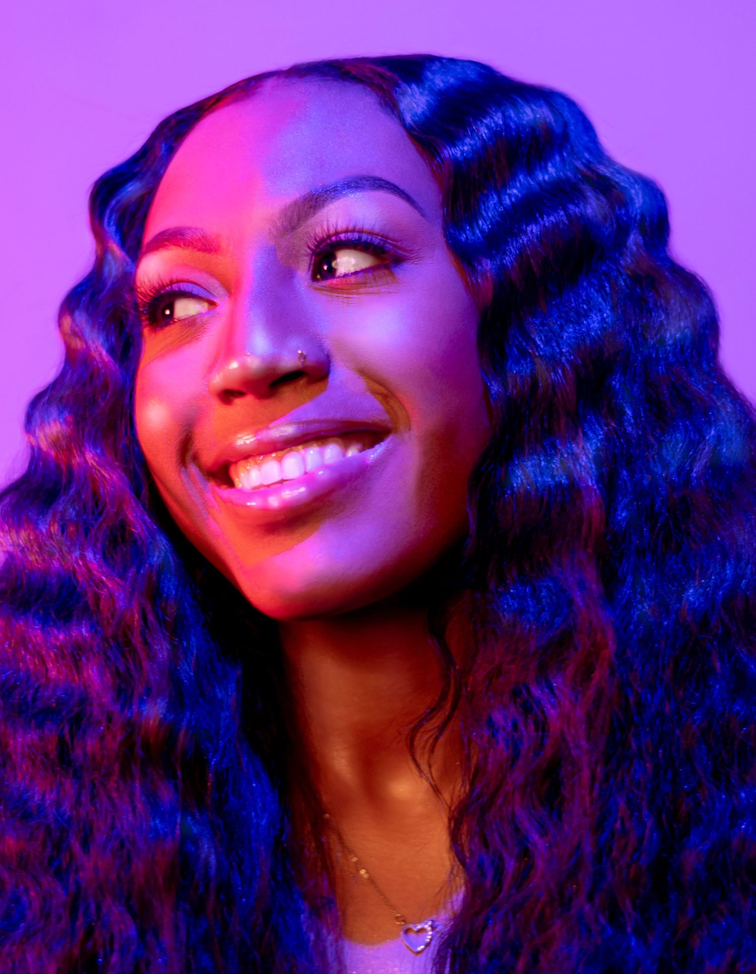 Artistic portrait of a smiling woman with wavy hair under vibrant purple and pink lighting.