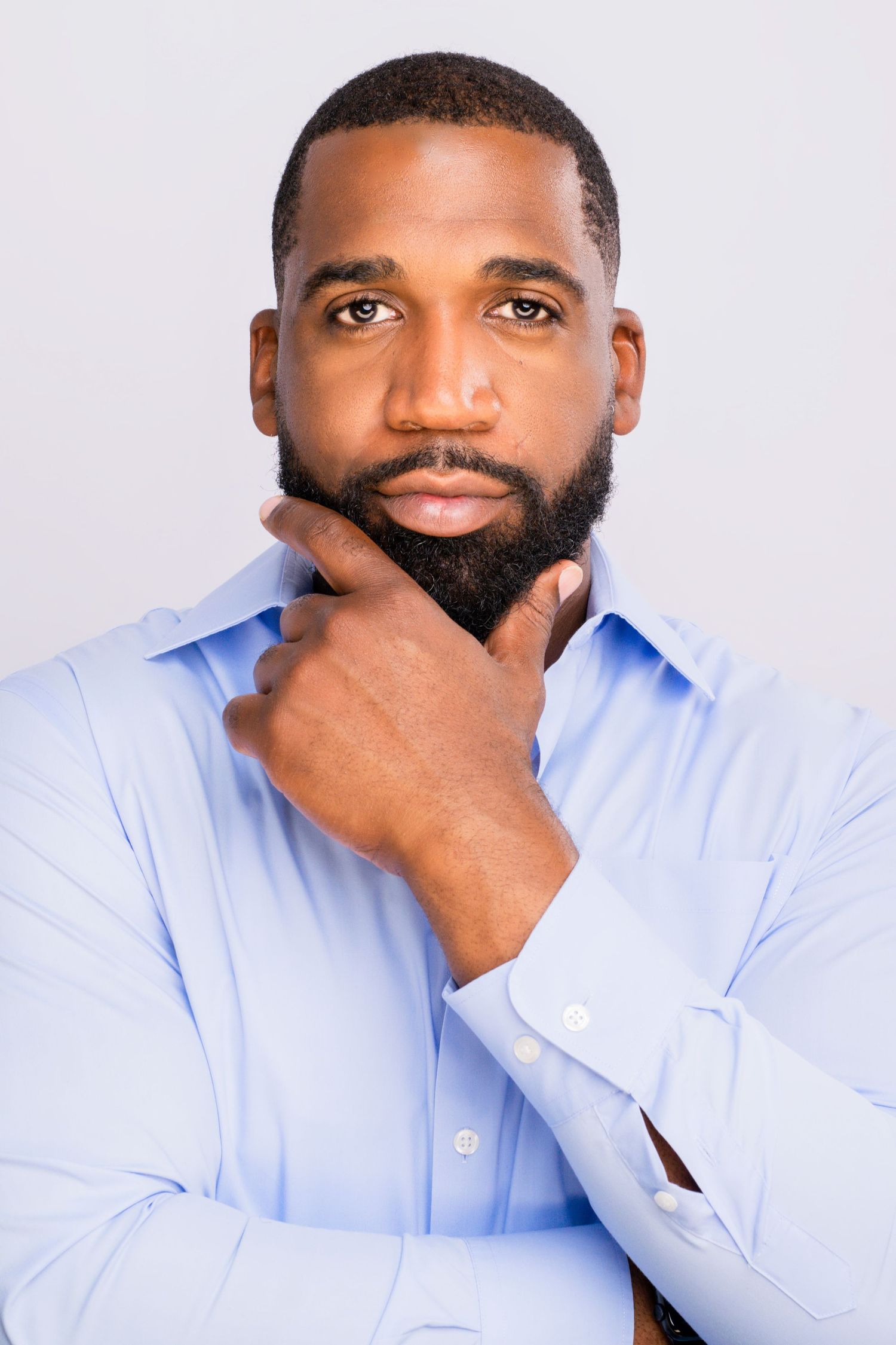 Confident Black man in a blue dress shirt touching his beard against a white background.