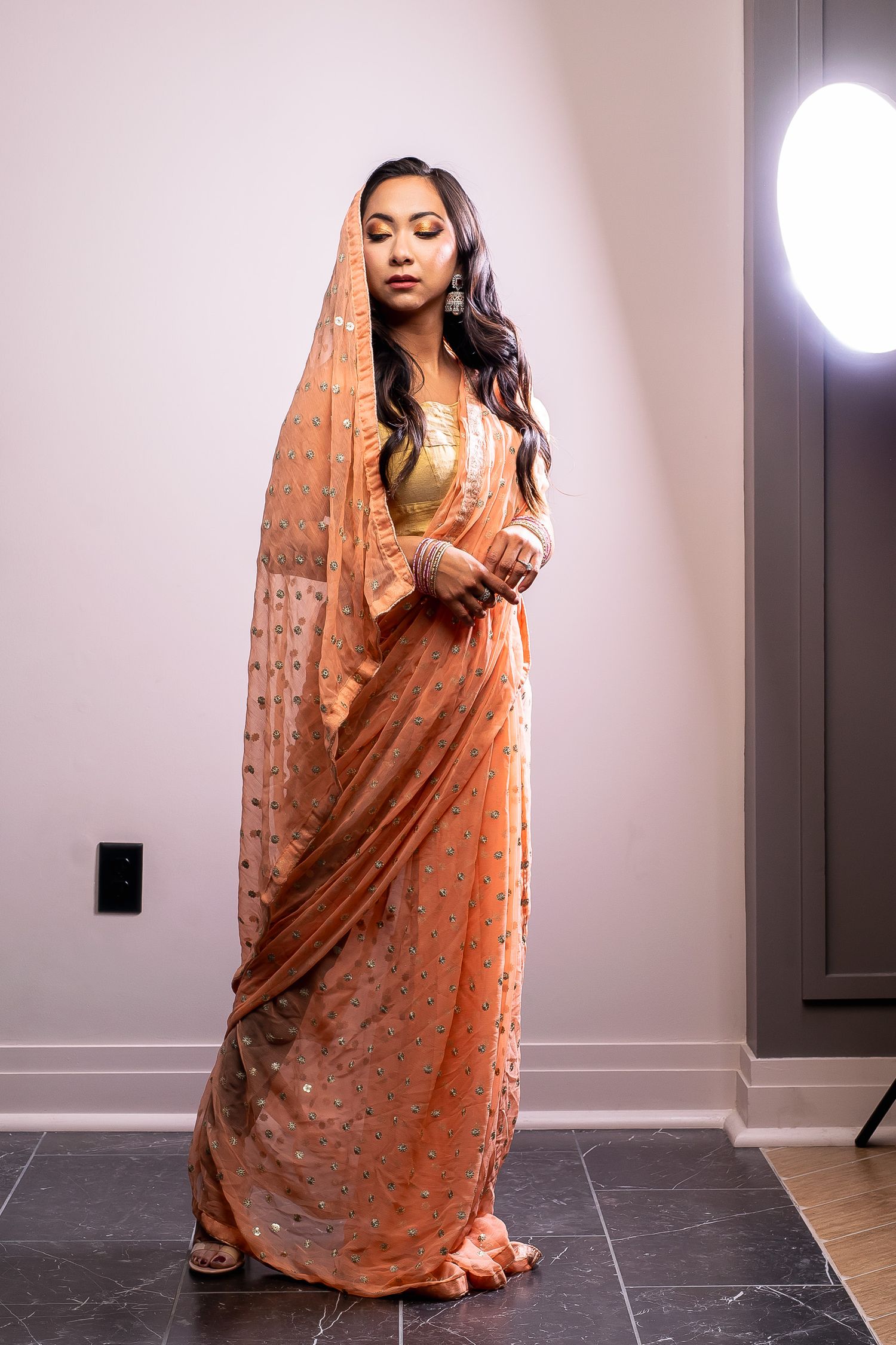 Woman in a peach embroidered saree with dupatta draped over her head posing in a softly lit room.