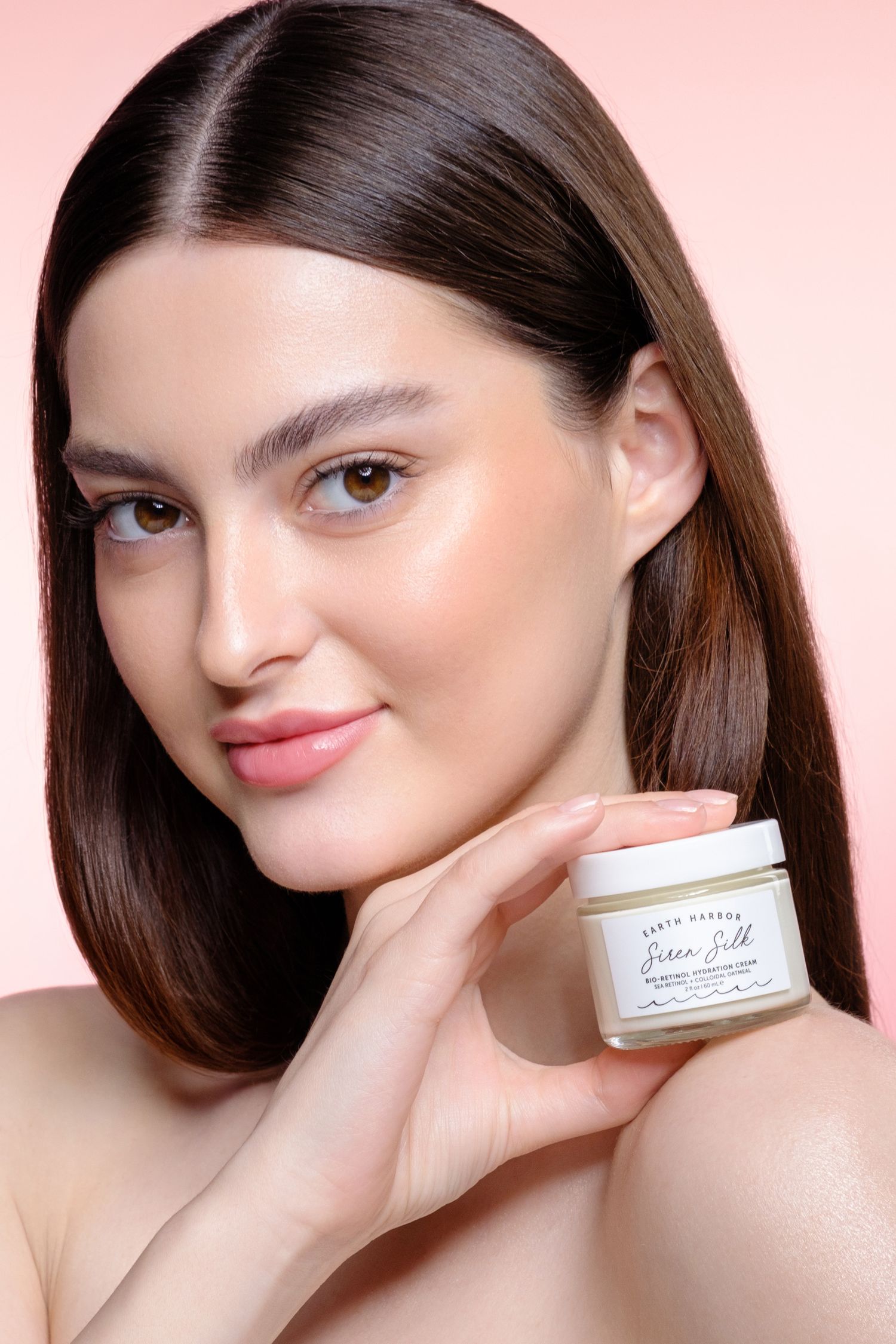 Young woman with glowing skin holding an Earth Harbor Siren Silk face cream jar against a pink gradient background.