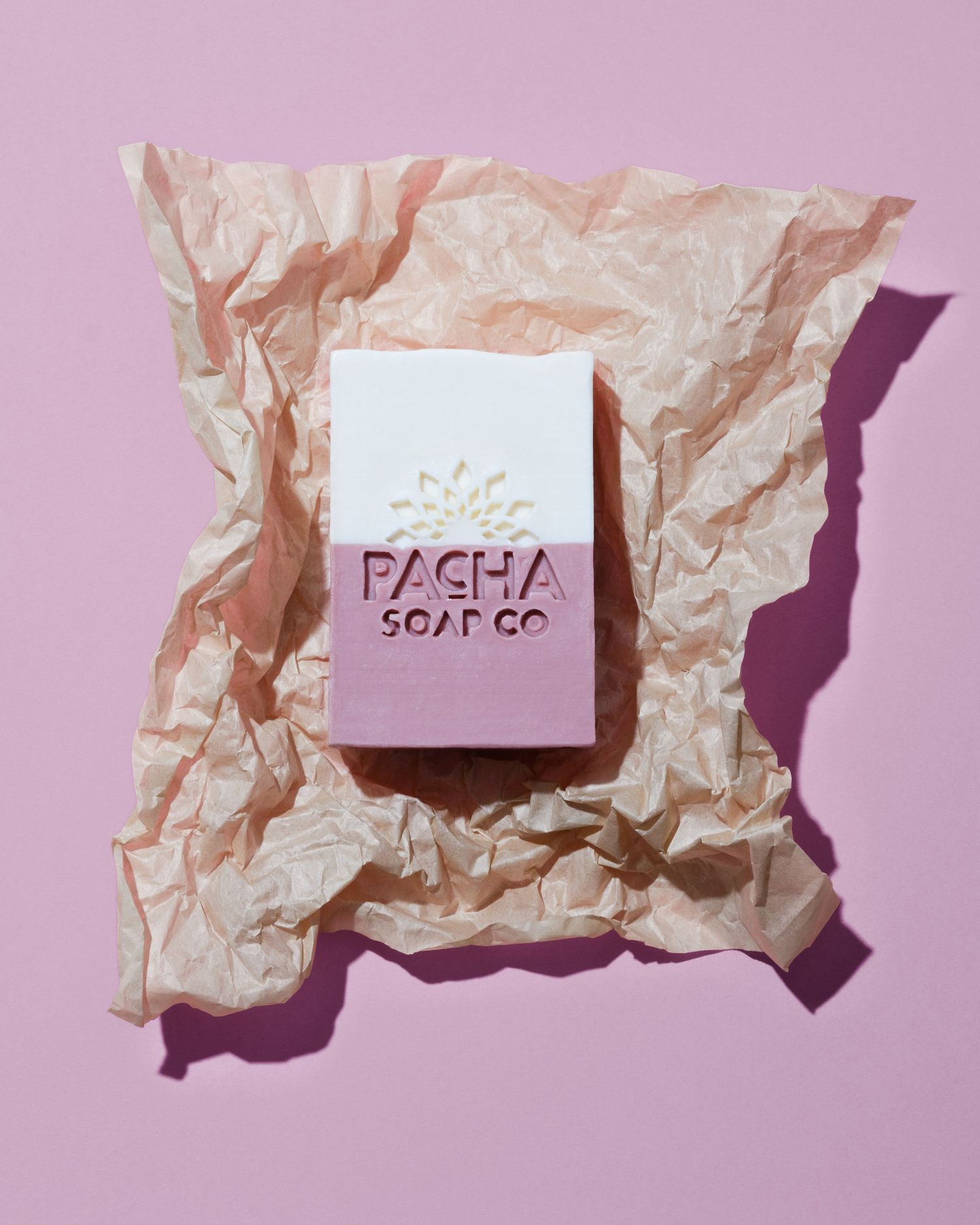 Handcrafted duo tone Pacha soap bar against beige crumpled paper casting a shadow against a light pink background, clean beauty product.