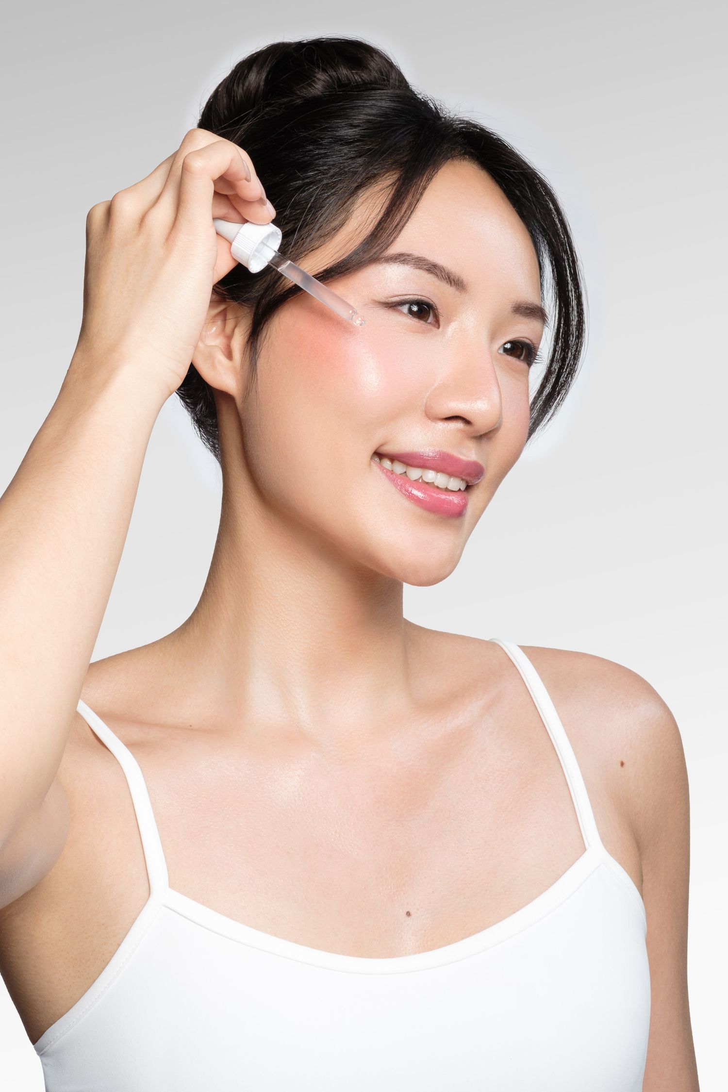 Young Asian woman applying facial serum with dropper, wearing white tank top, smiling against light gray background.