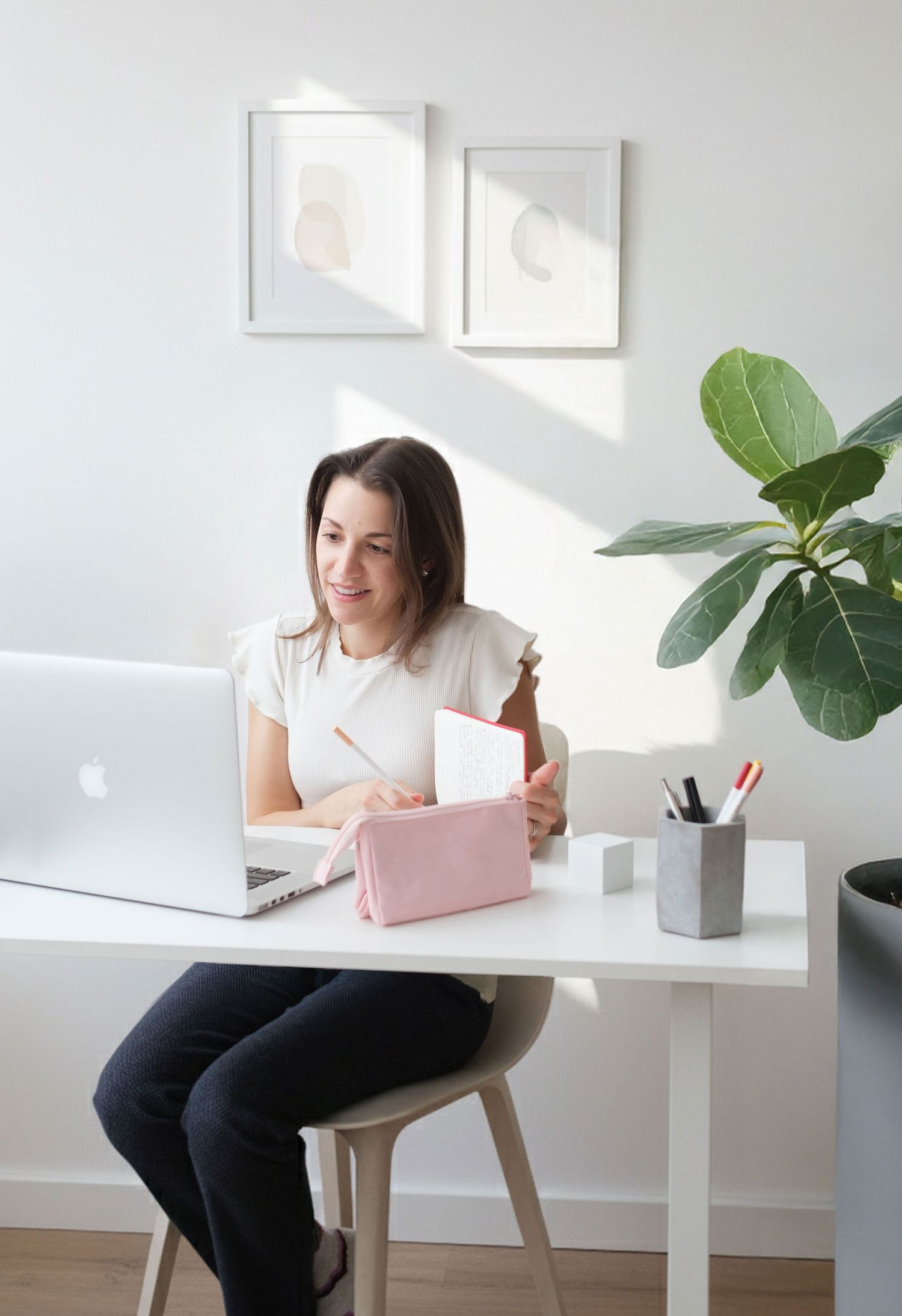 Darina sitting at her desk working on her laptop, holding a pen and notebook in a bright minimalist home decor.