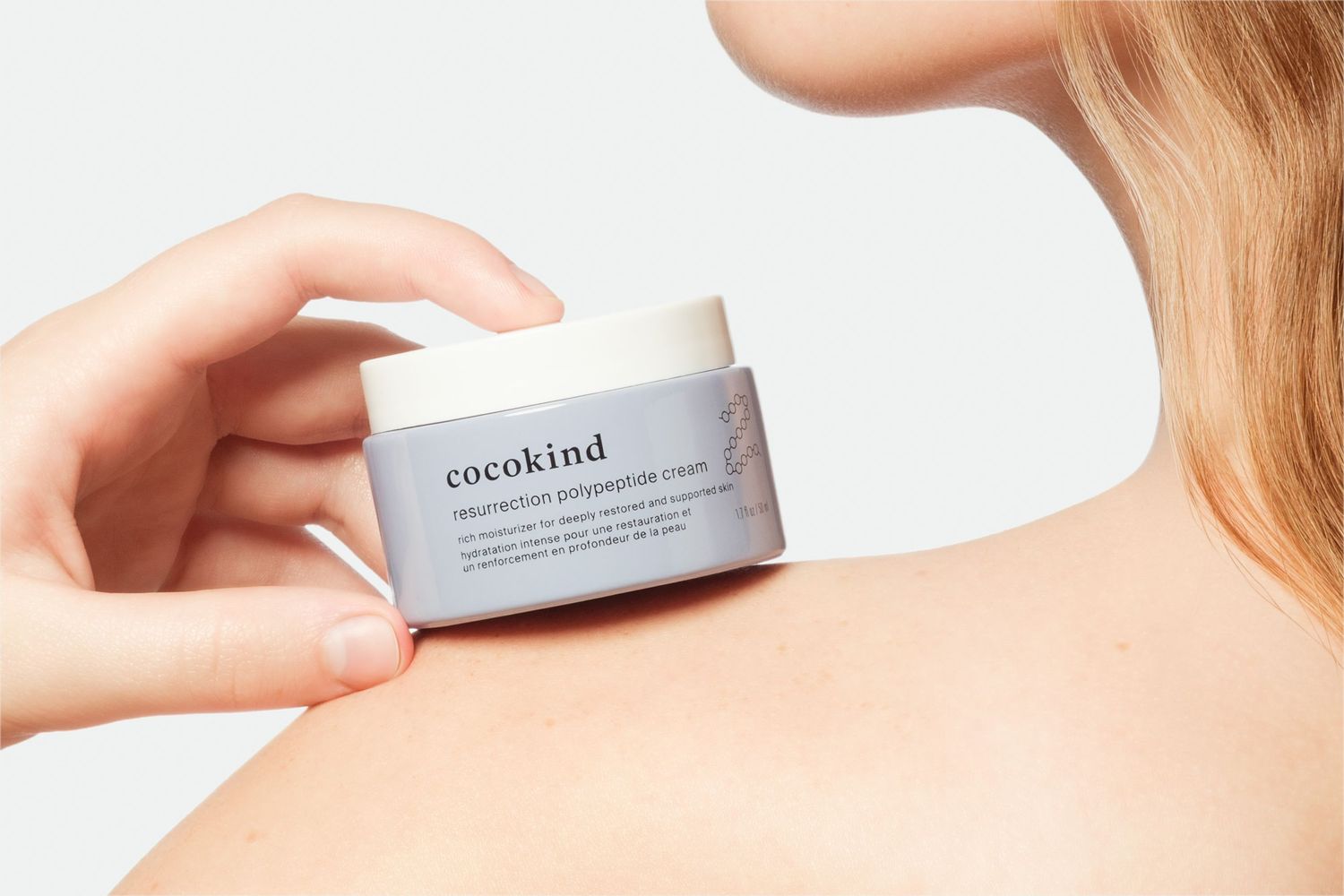 Hand holding Cocokind moisturizing cream jar against a light background near the woman's shoulder.