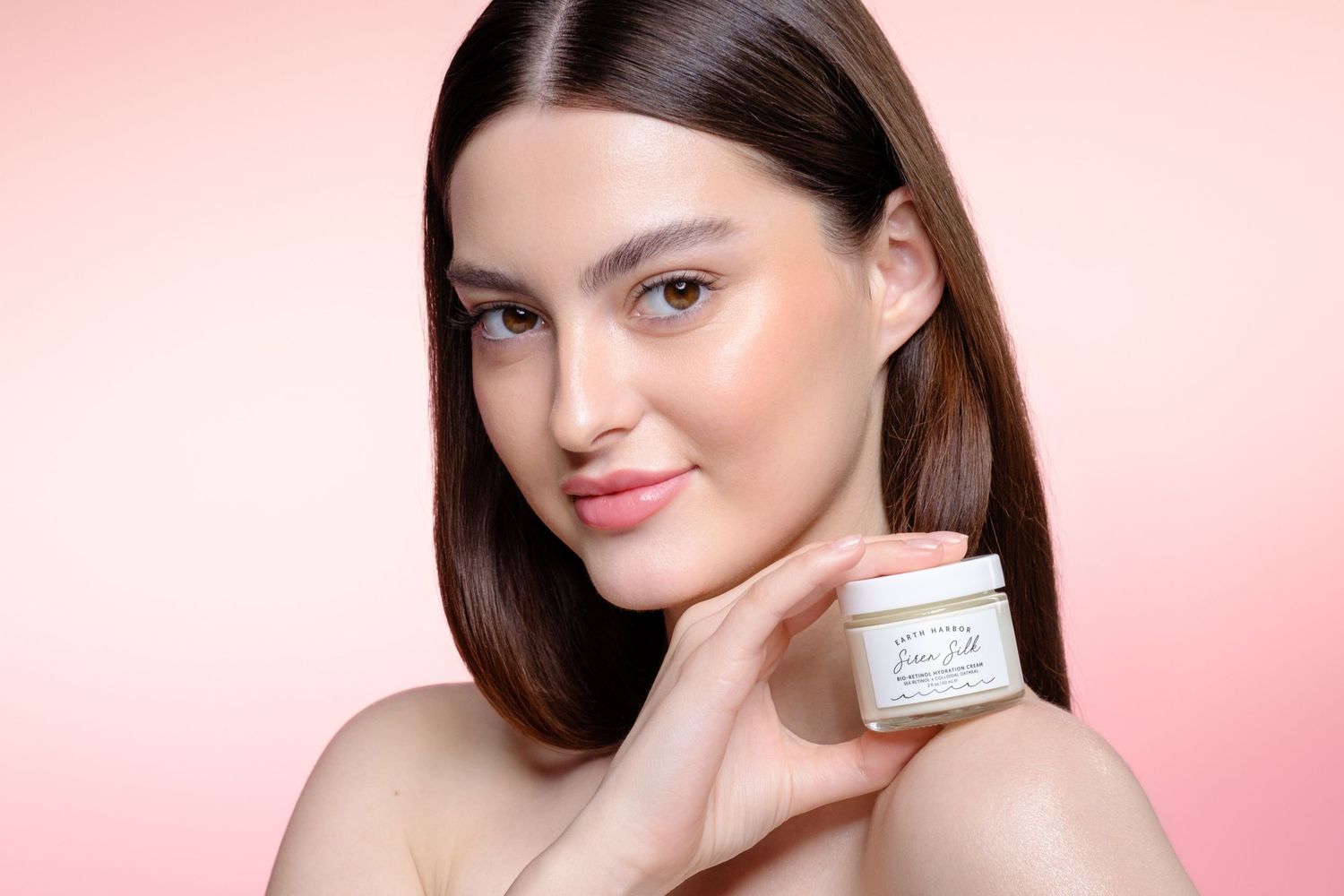 Young woman with glowing skin holding an Earth Harbor Siren Silk face cream jar against a pink gradient background.