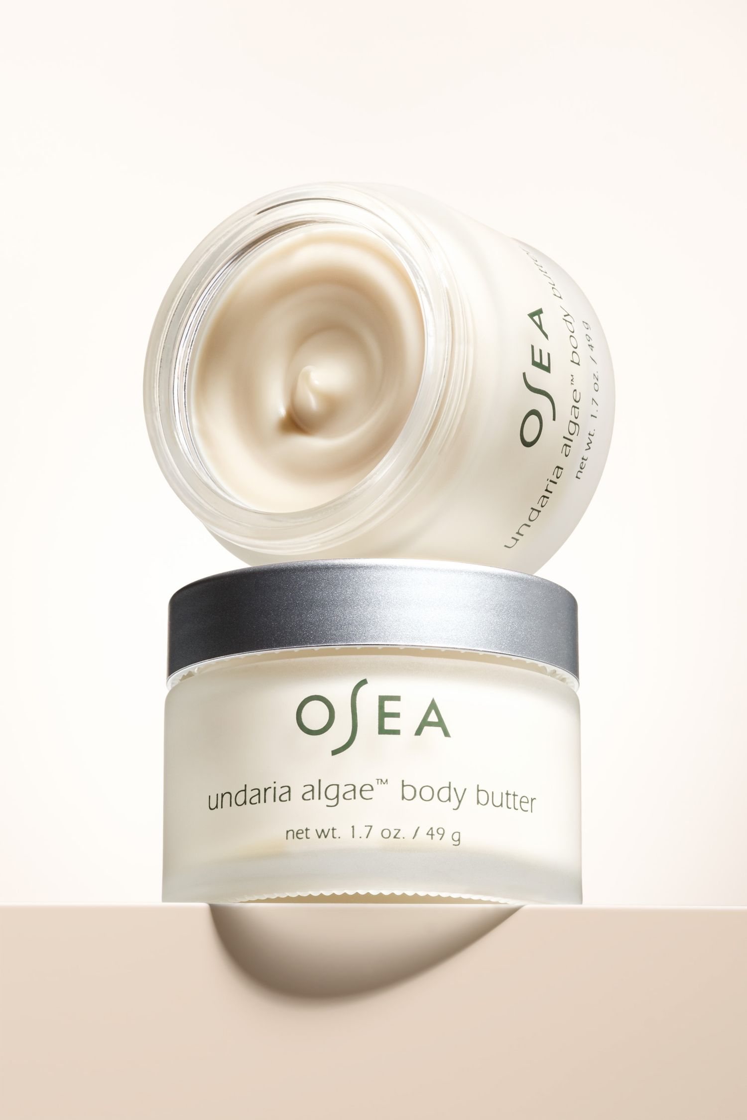 Two Osea body butter jars, stacked against neutral background, one open showing the creamy texture, professional studio product photography.