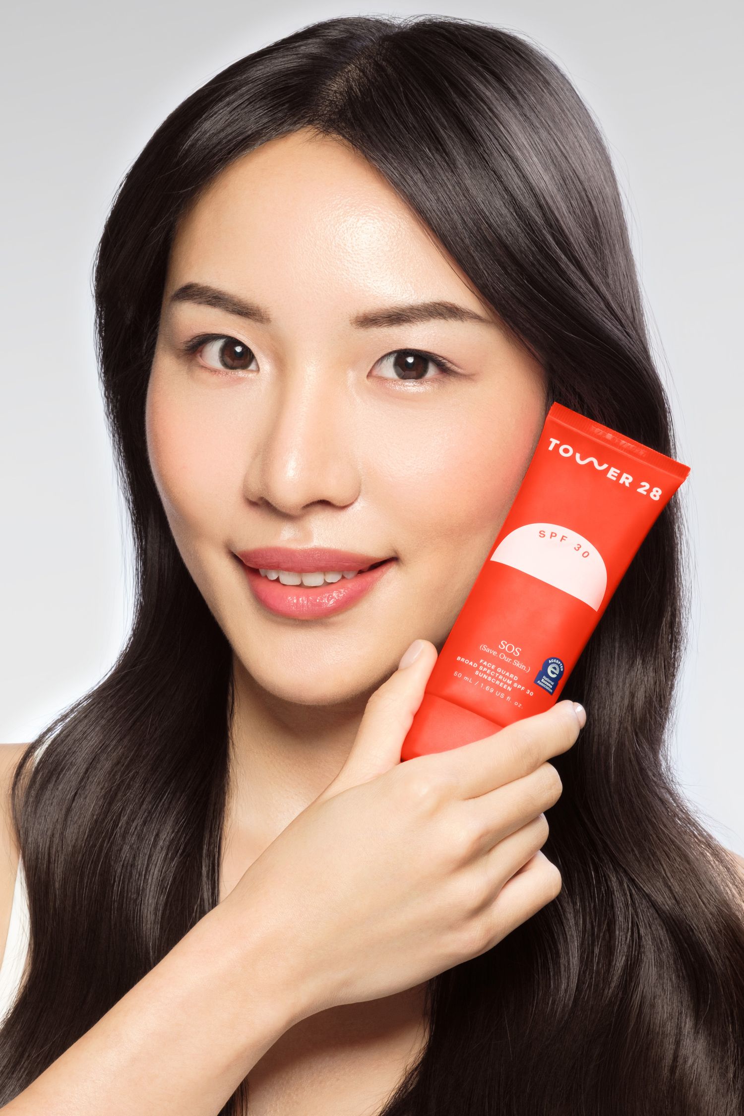 Professional studio photo of an asian woman holding Tower 28 SPF 30 sunscreen tube against her cheek, showcasing the red product packaging.