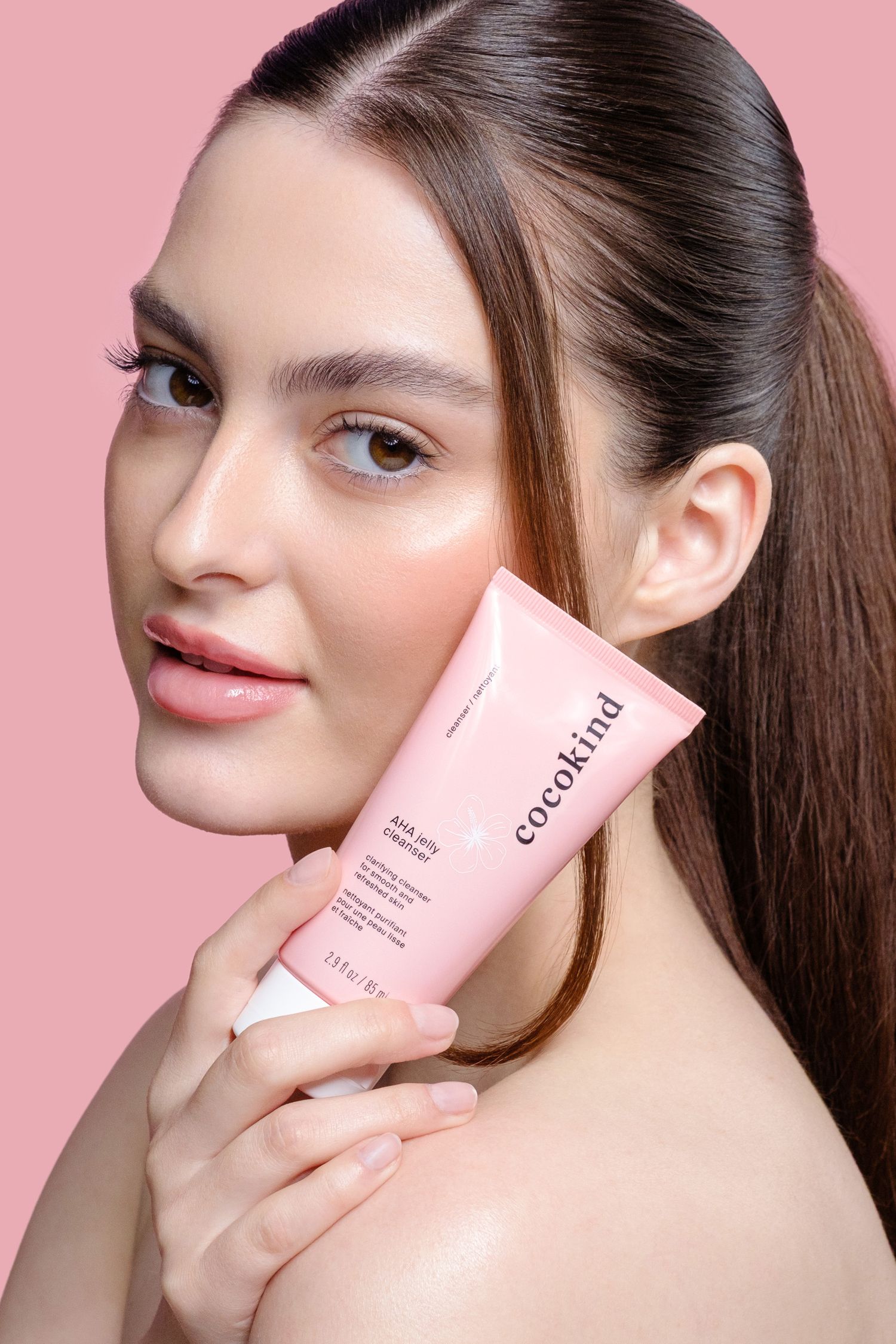 Young woman with glowing skin holding a pink Cocokind AHA Jelly Cleanser tube against her cheek on a pink background. Professional studio.