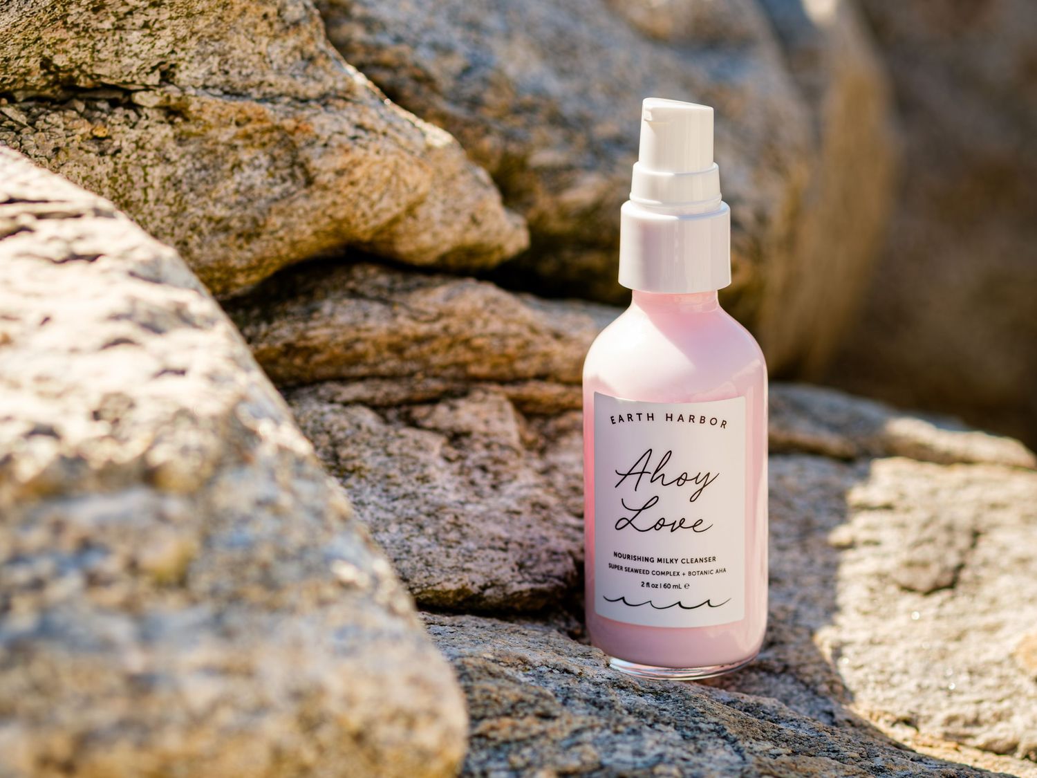 Earth Harbor Ahoy Love cleanser resting on rocky coastal stones, true beach scene with strong sunlight illuminating the top of the bottle.