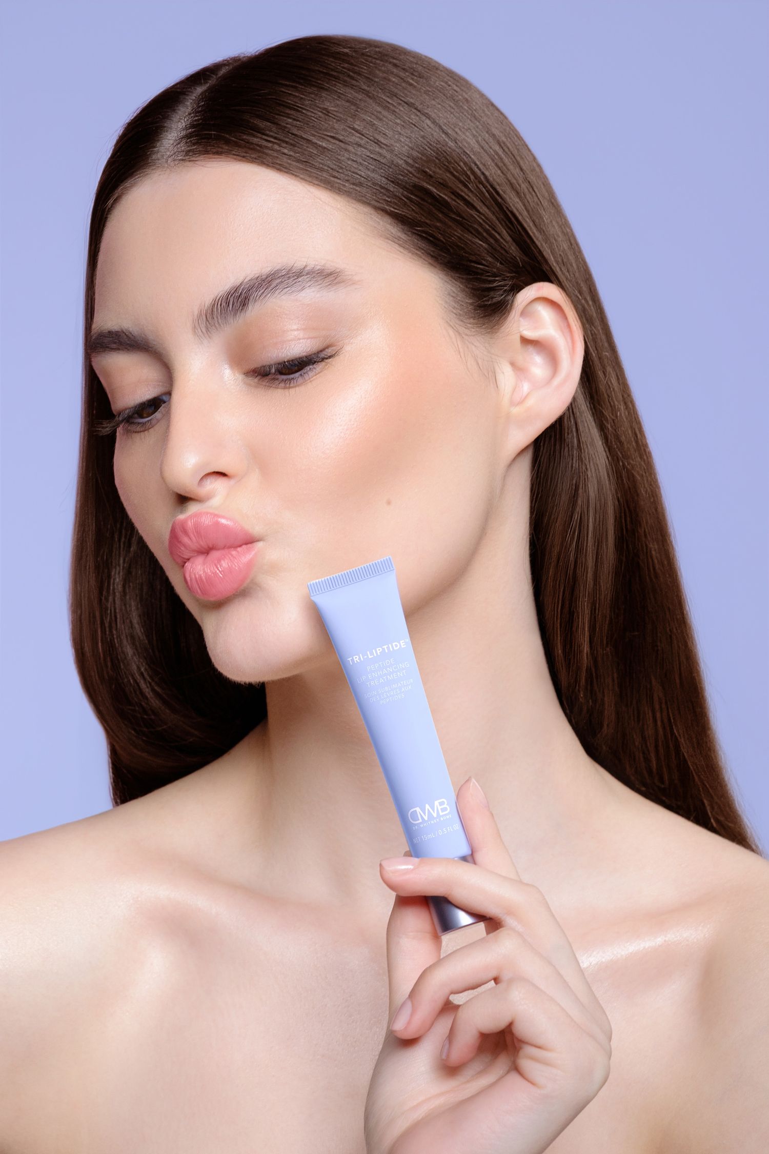 Young woman with glowing skin holding a tri-liptide lip enhancing treatment with her lips puckering on a purple background.