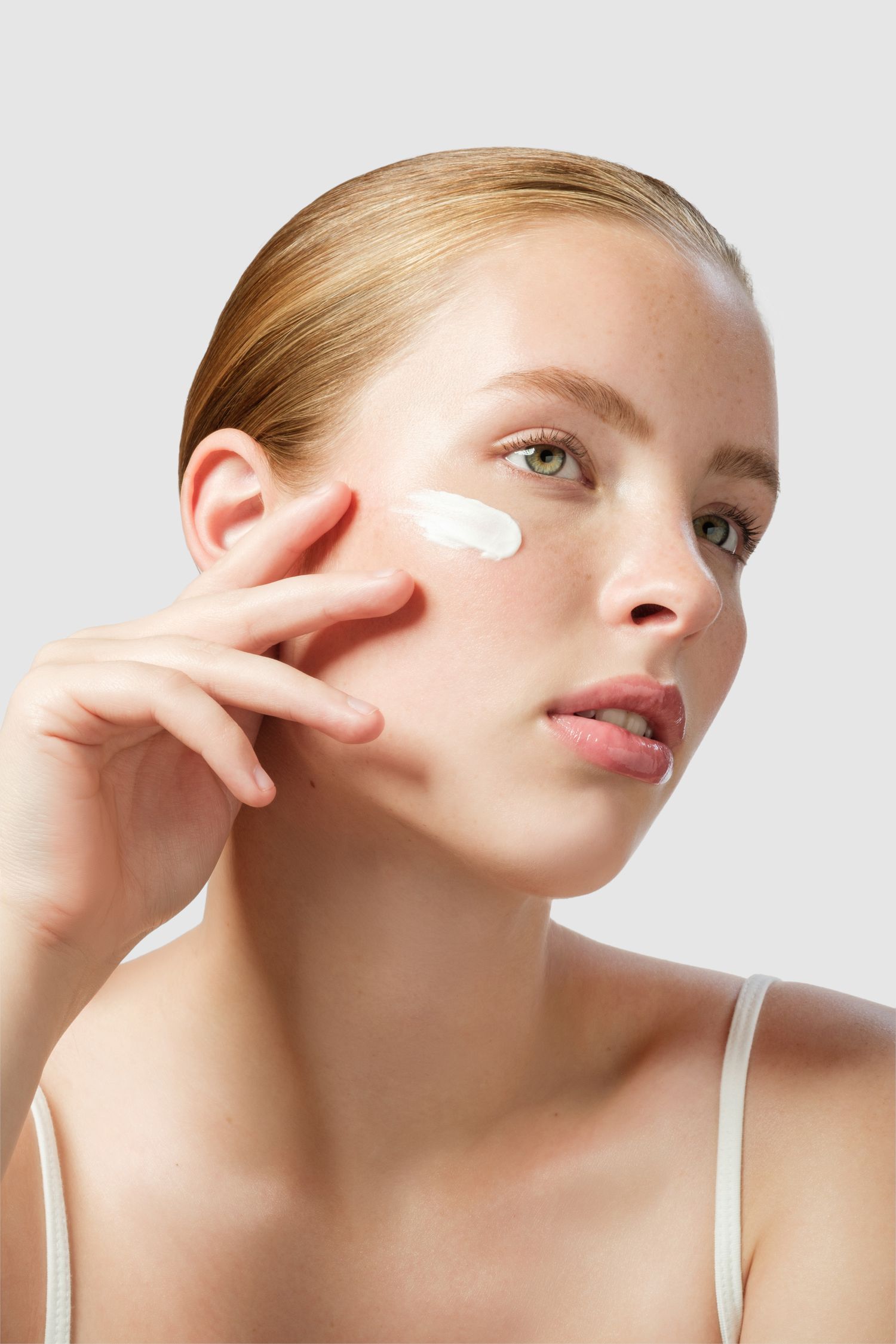Young woman with flawless skin applying white face cream to her cheek against a light gray background. Professional studio beauty image.