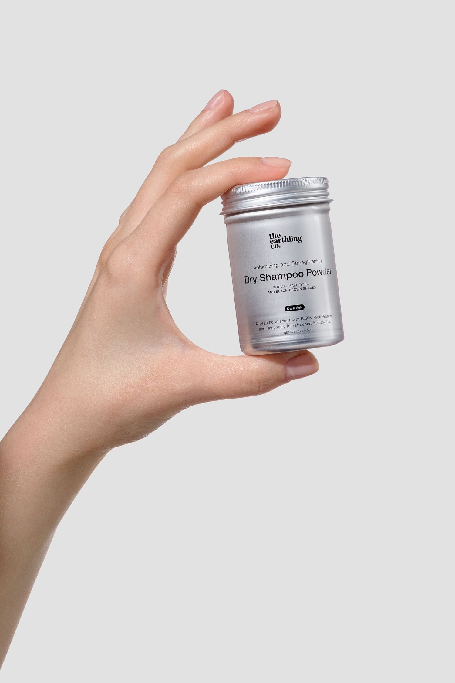 Woman's hand holding a The Earthling Co small silver jar of dry shampoo powder against a light gray background. Professional studio photo.