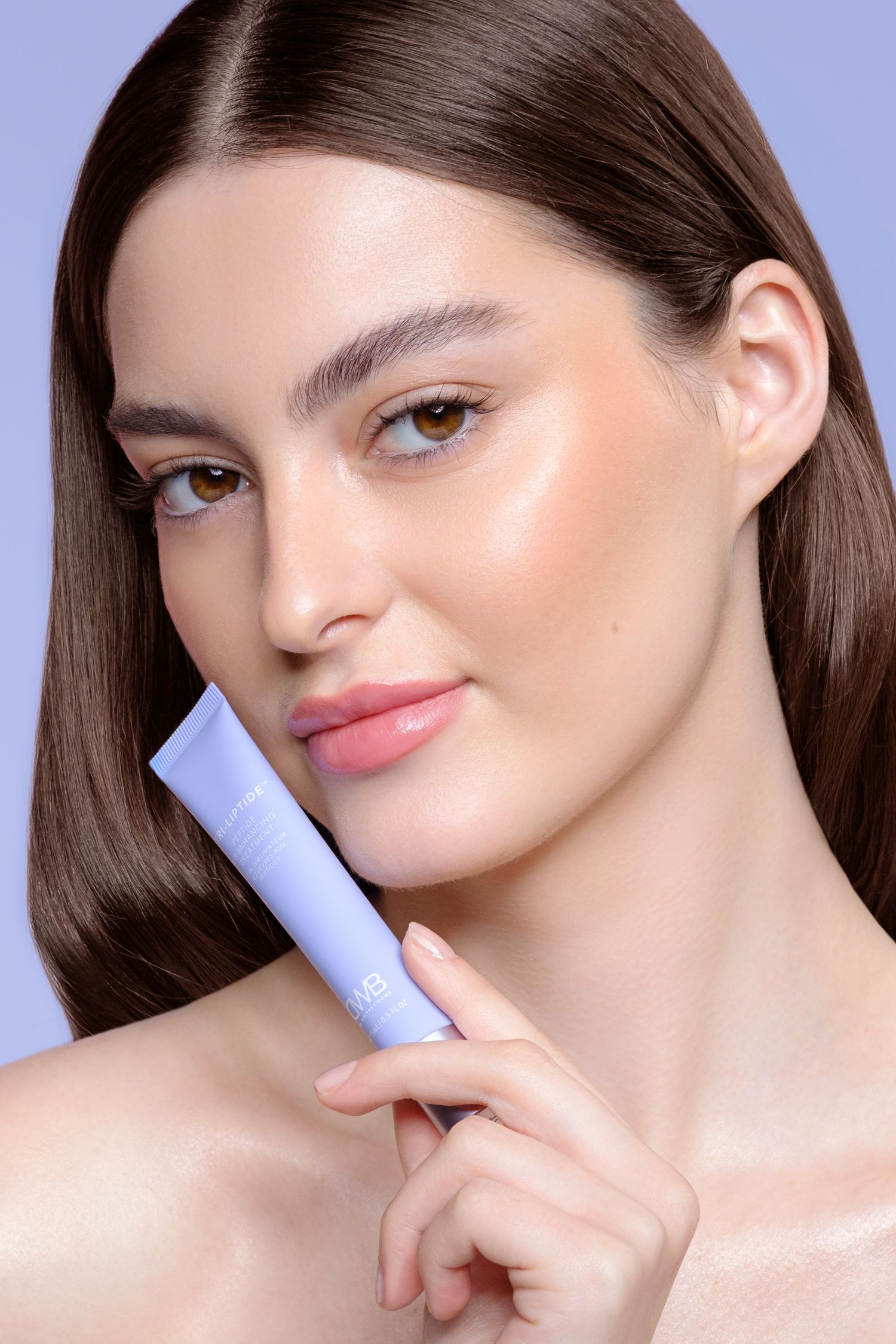 Young woman with glowing skin holding a tri-liptide lip enhancing treatment against her cheek on a purple background.